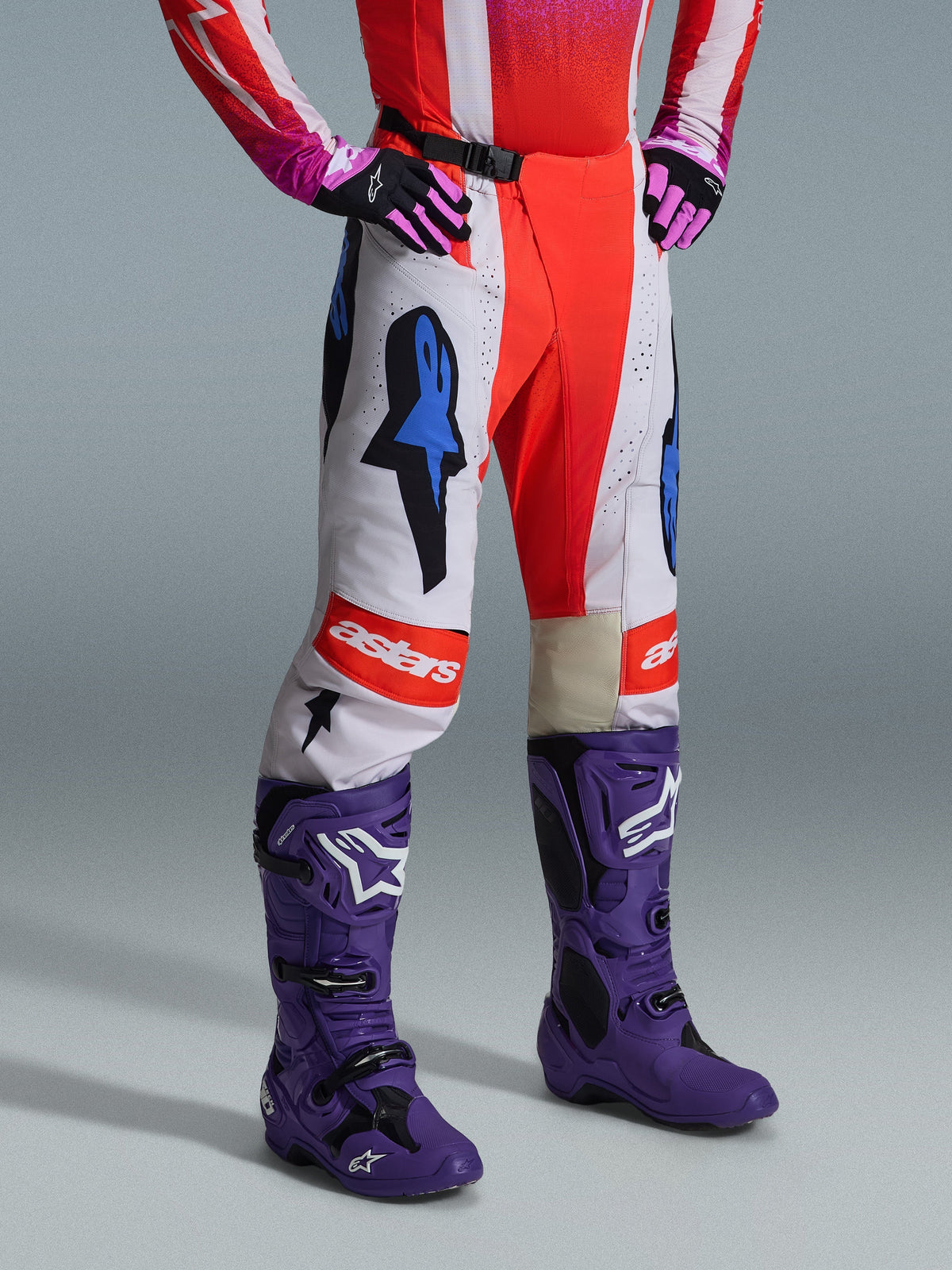 Alpinestars 2026 Techstar Knif Pants, pantaloni da motocross, bianco e arancione con accenti blu, indossati come parte di un set completo con maglia rossa e rosa, stivali viola e guanti rosa, con loghi vistosi e vestibilità racing atletica