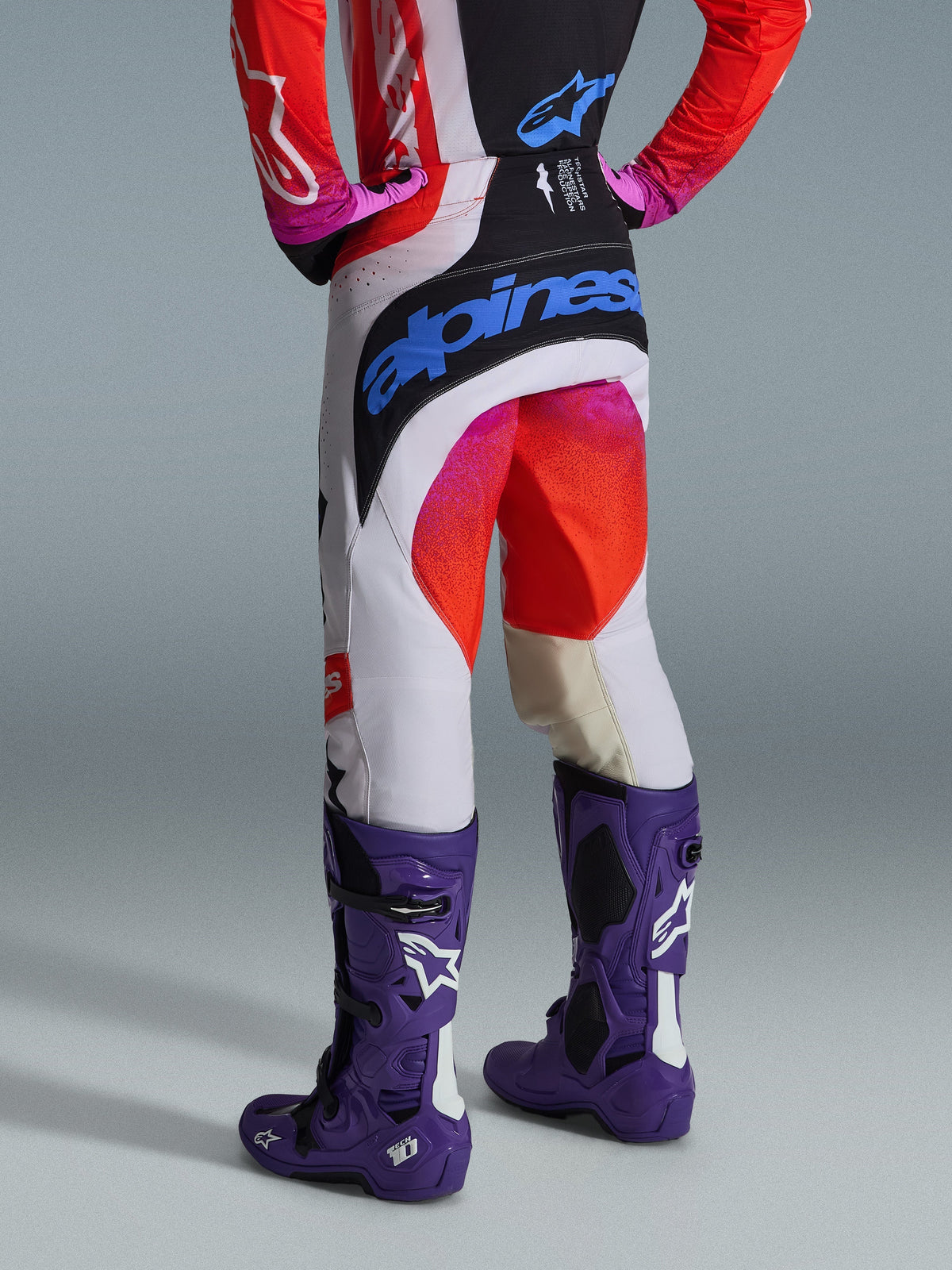 Alpinestars 2026 Techstar Knif Pants e Gear Set, vista posteriore di un pilota di motocross, pantaloni bianchi con pannelli neri, accenti sfumati rosa e rossi, grande logo Alpinestars blu sulla seduta, stivali Tech 10 viola e maglia coordinata rossa e nera