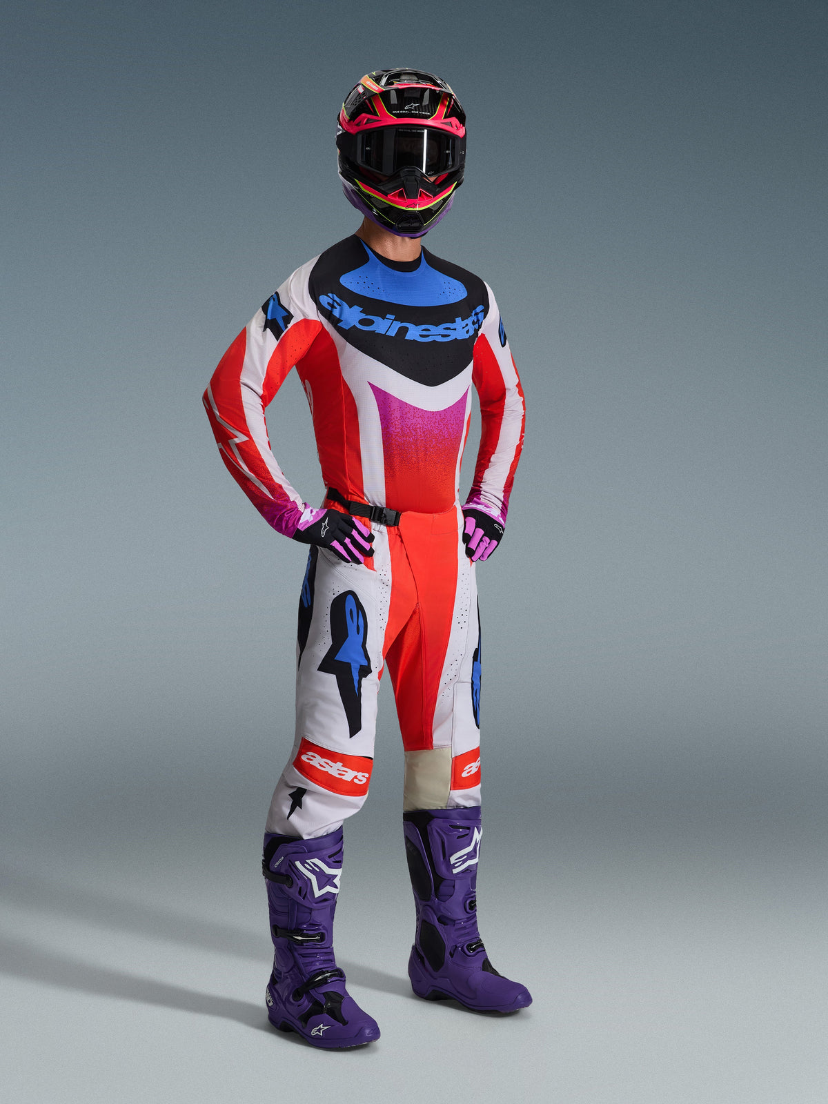 Pilota di motocross con abbigliamento racing completo Alpinestars, maglia Techstar Knif 2026 bianca e rossa con accenti blu e rosa, pantaloni da motocross coordinati, stivali viola e casco multicolore, outfit professionale coordinato per prestazioni off-road