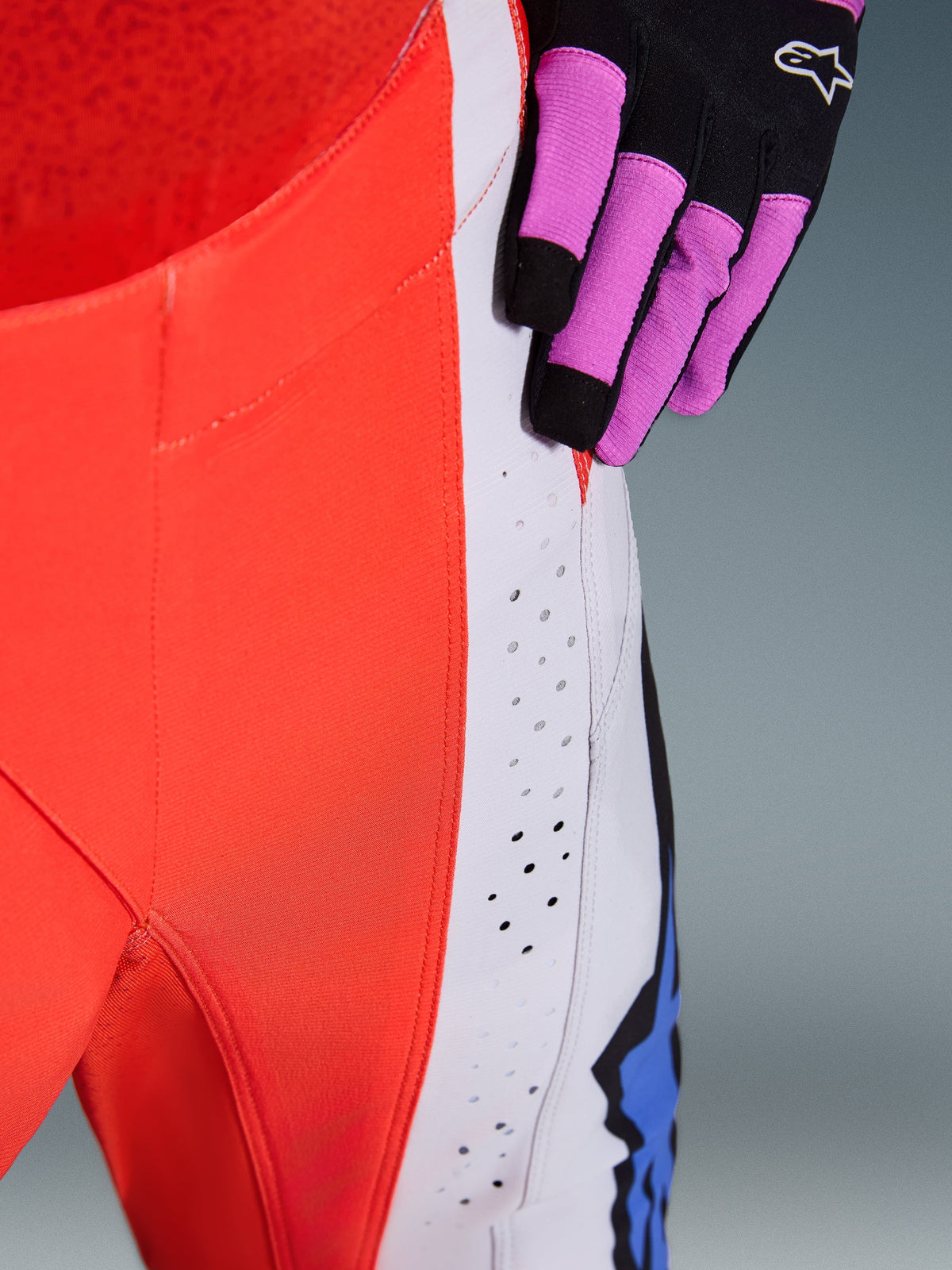 Pantaloni Alpinestars Techstar Knif 2026, primo piano del fianco e del pannello laterale in Arancione Fluo e Grigio Chiaro, con perforazioni al laser per il flusso d'aria, indossati con un guanto da motocross nero e viola, abbigliamento racing ad alte prestazioni