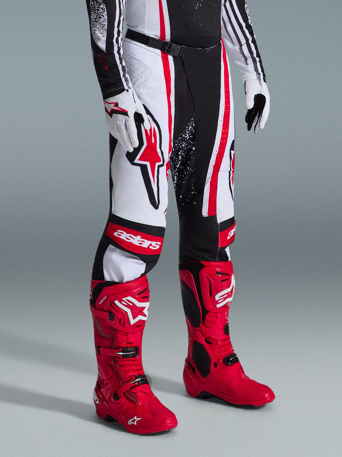 Alpinestars 2026 Techstar Nomur Pants, pantaloni da motocross, nero, bianco e rosso, indossati con vivaci stivali da corsa rossi e guanti bianchi, con audaci strisce verticali, un grande logo a stella rossa sulla coscia e pannelli rinforzati sulle ginocchia per prestazioni off-road.