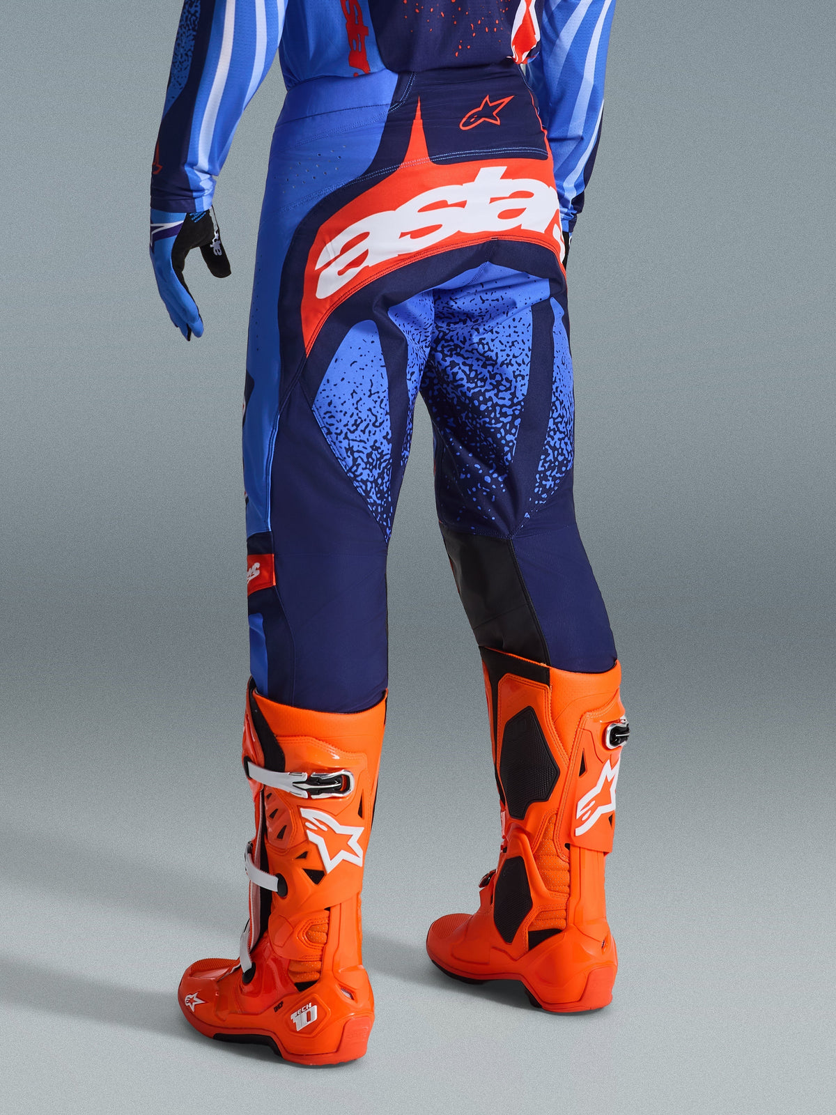 Alpinestars 2026 Techstar Nomur Pants, pantaloni da motocross, Dark Navy, arancione e blu UCLA, vista posteriore con logo "Astars" bianco sulla seduta, indossati con stivali da motocross arancione acceso e maglia coordinata, vestibilità atletica per gare fuoristrada