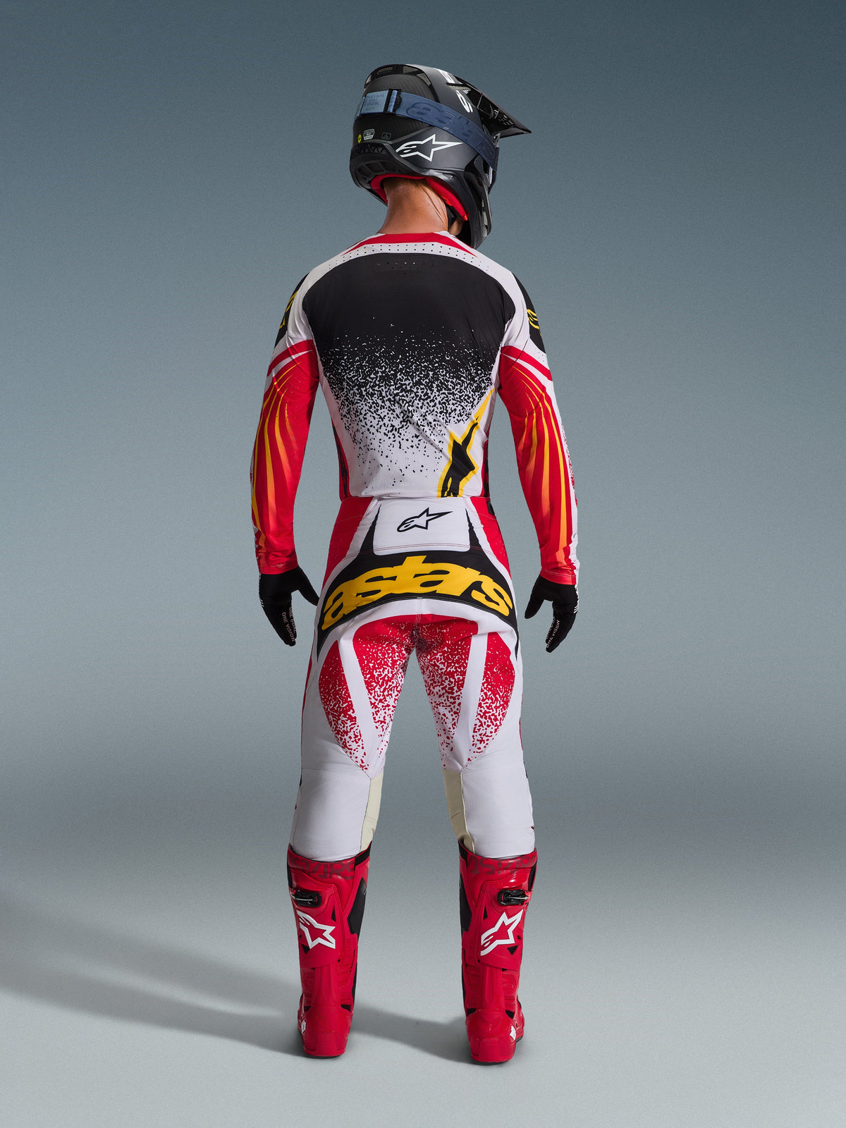 Completo Alpinestars Techstar Nomur 2026, abbigliamento da motocross visto dal retro, maglia bianca con effetto splatter nero e maniche con grafiche rosso-arancio, pantaloni bianchi e rossi con logo 'astars' giallo, stivali da corsa rossi, casco nero con trama in carbonio e occhiali blu, equipaggiamento off-road ad alte prestazioni