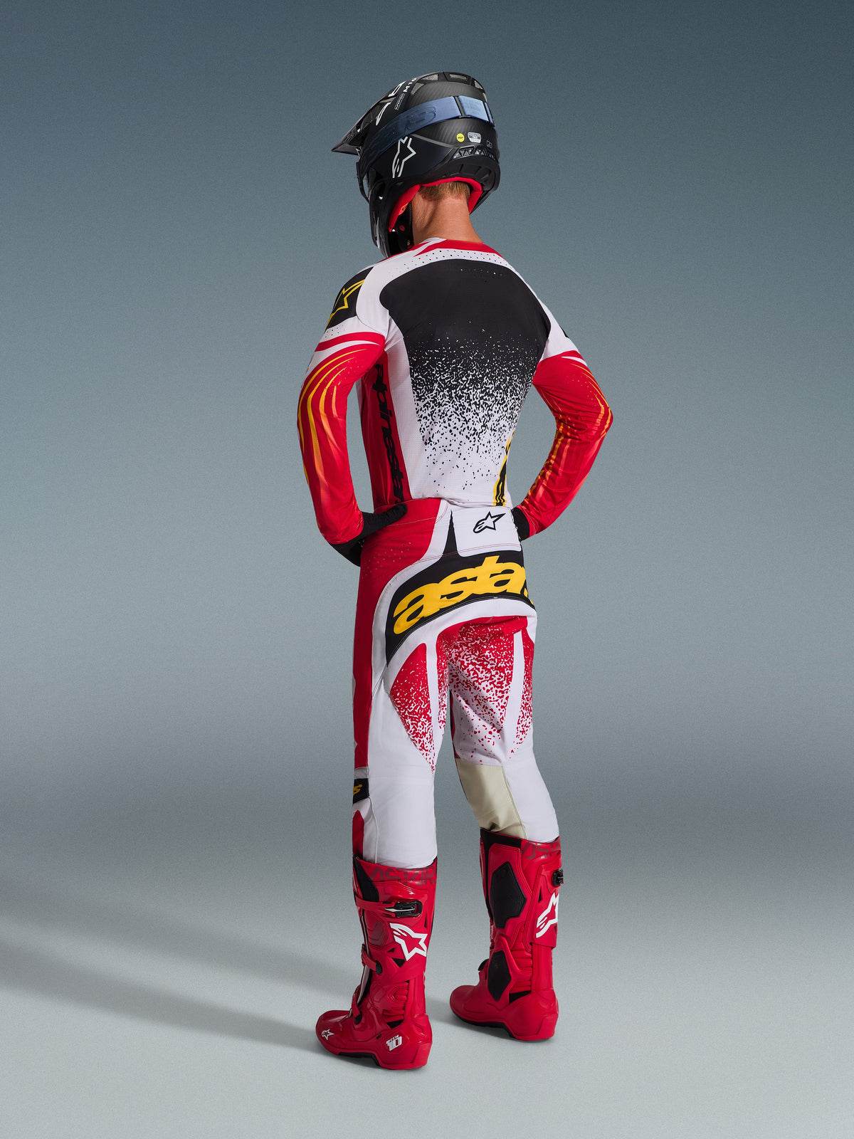 Pilota di motocross visto da dietro che indossa il completo Alpinestars 2026 Techstar Nomur, maglia rossa, bianca e nera con schiena bianca puntinata, pantaloni coordinati con logo 'astars' giallo sulla seduta, stivali da corsa rosso acceso e casco nero, outfit completo da motocross coordinato