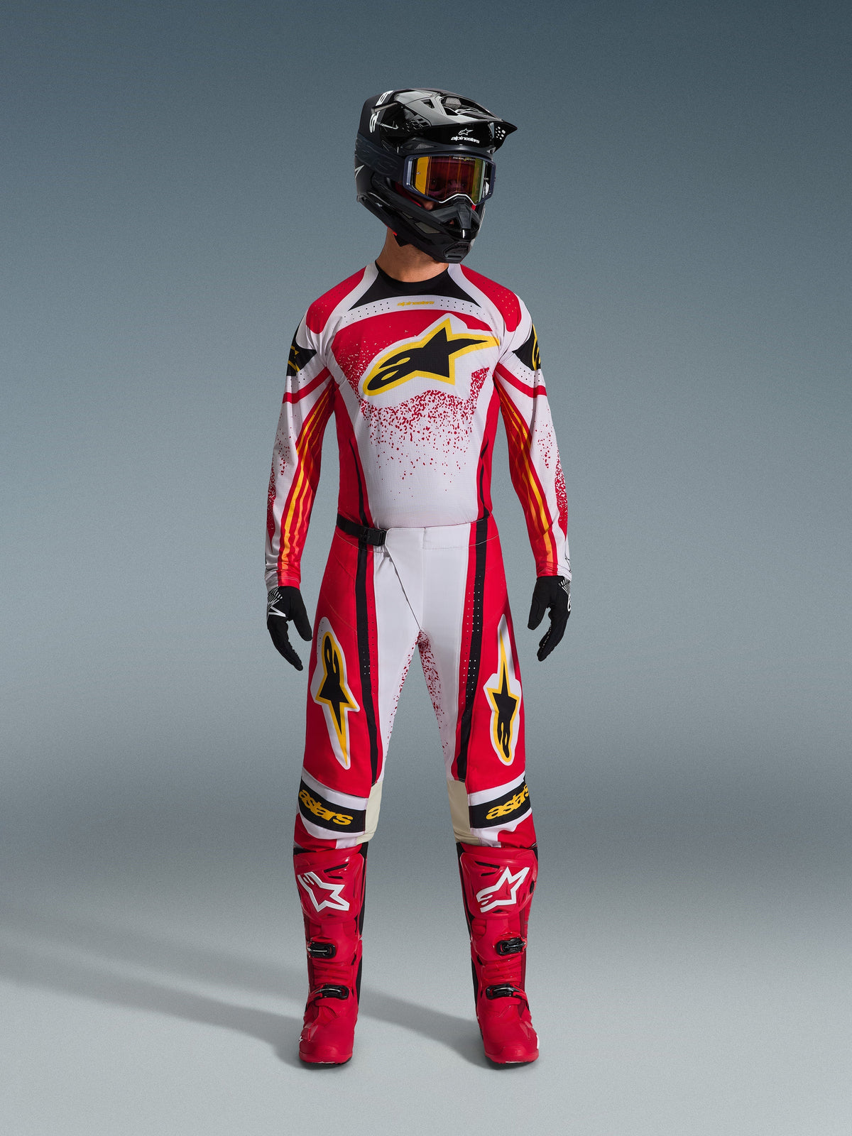 Alpinestars Techstar Nomur Jersey 2026, maglia da motocross rossa e bianca con accenti neri e loghi gialli, indossata da un pilota con pantaloni abbinati, stivali rossi e casco nero, abbigliamento professionale per corse fuoristrada