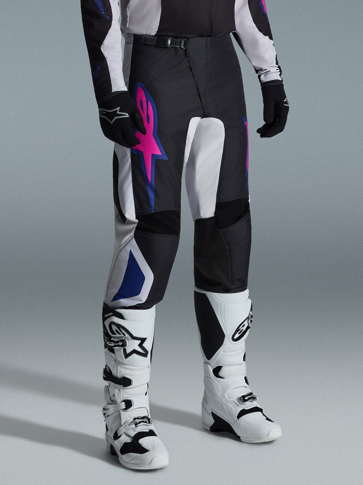 Alpinestars 2026 Fluid Grid Pants, pantaloni da motocross, nero, grigio chiaro e viola, con logo a stella rosa e accenti geometrici blu, indossati con stivali da motocross bianchi e guanti neri, abbigliamento ergonomico per la guida fuoristrada