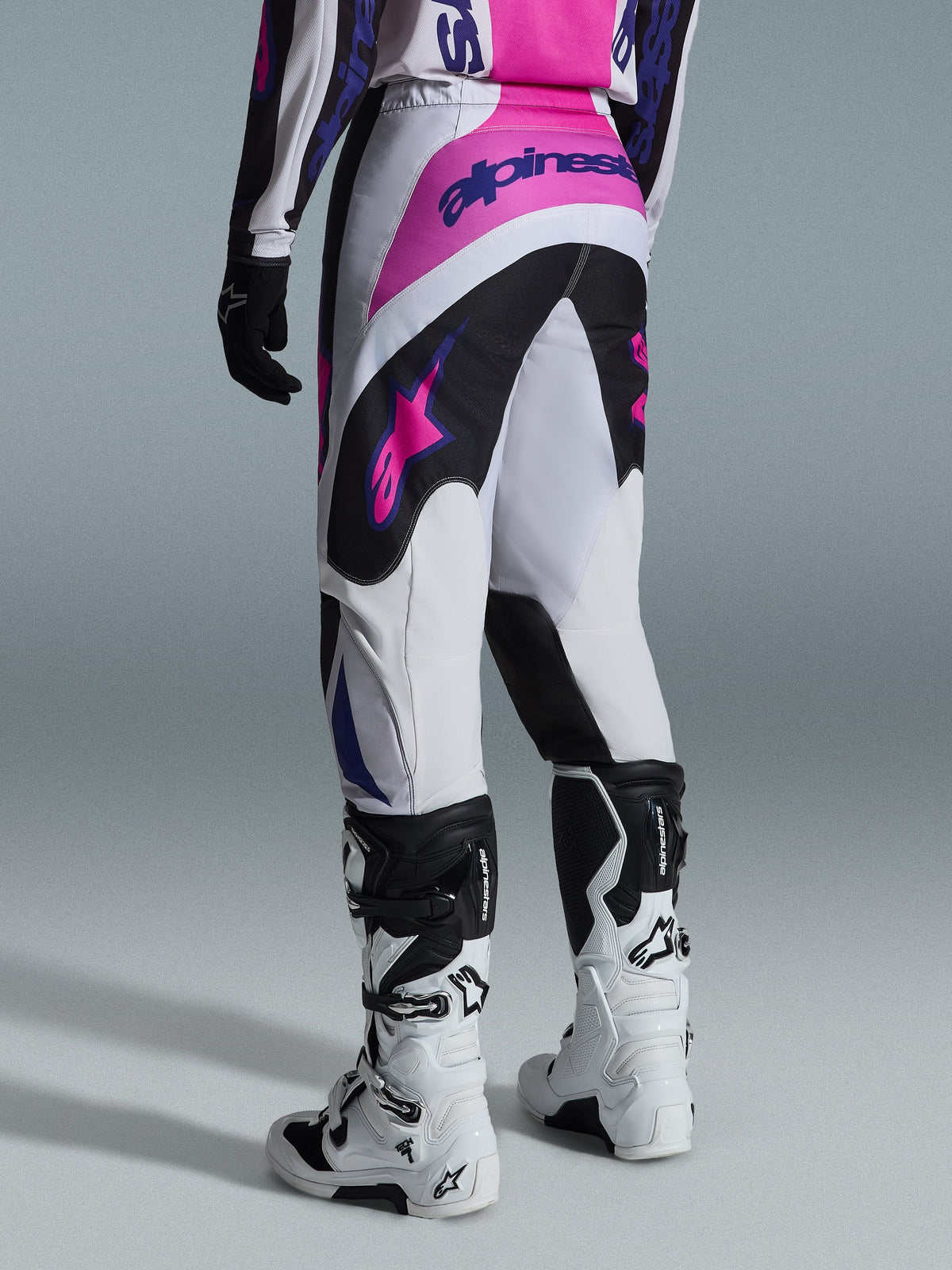 Alpinestars 2026 Fluid Grid Pants, pantaloni da motocross, nero, grigio chiaro e viola, vista posteriore del pilota con stivali bianchi e neri, loghi Alpinestars rosa e viola, vestibilità atletica per corse fuoristrada