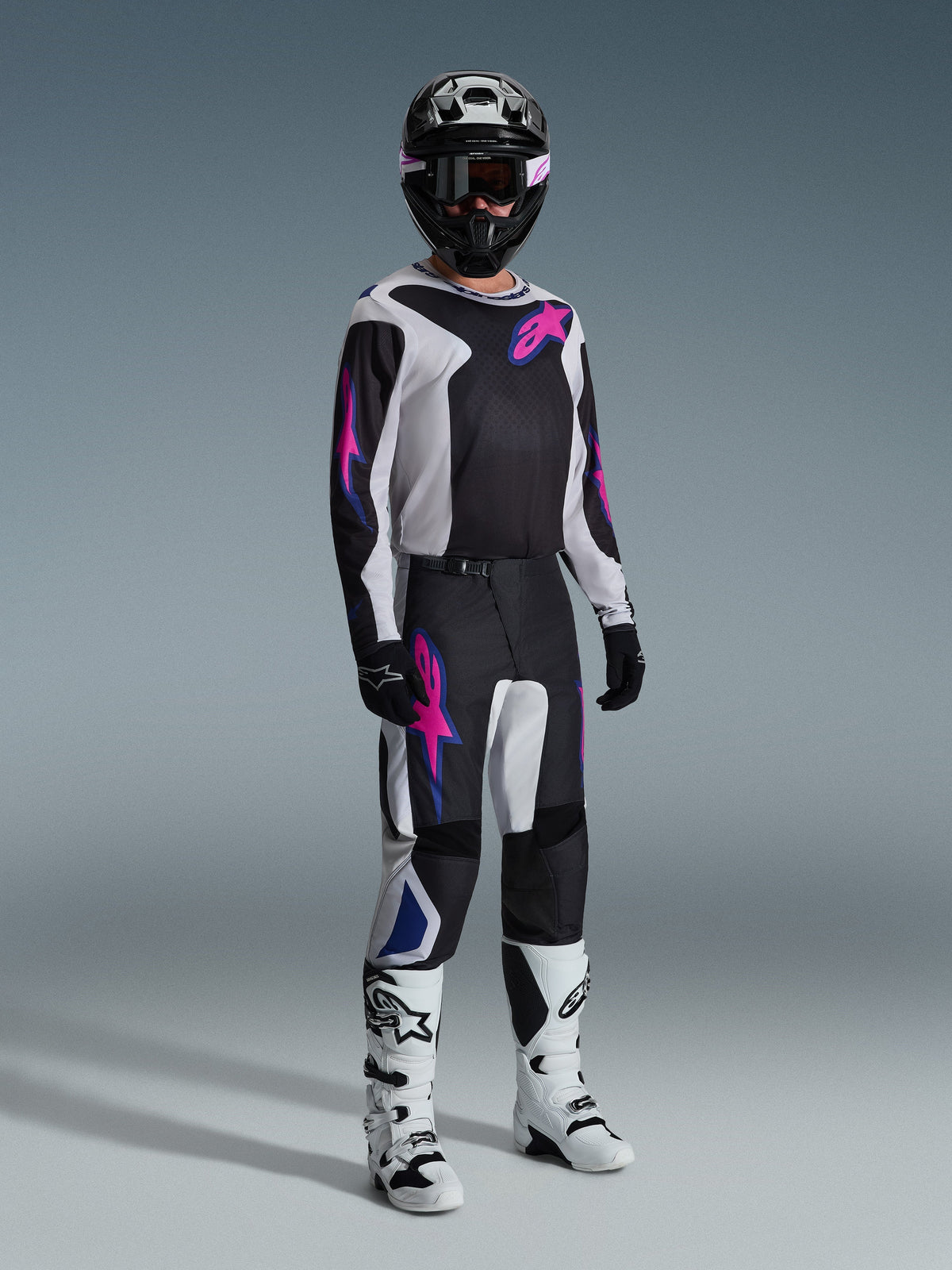 Pilota di motocross con abbigliamento completo da gara, pantaloni Alpinestars 2026 Fluid Grid in Nero Grigio Chiaro e Viola, maglia coordinata con grafiche rosa e blu, casco e occhiali neri, stivali bianchi e neri e guanti neri, completo coordinato per corse fuoristrada