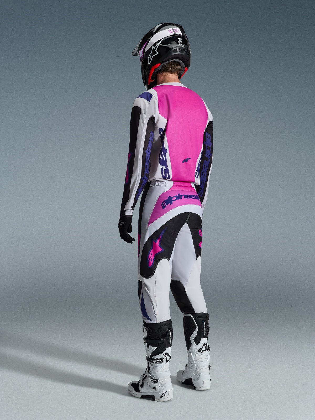 Alpinestars 2026 Fluid Grid Pants, pantaloni da motocross, nero, grigio chiaro e viola, vista posteriore del pilota con maglia coordinata e stivali bianchi, con loghi viola audaci e pannelli rosa vivaci, progettati per prestazioni motocross fuoristrada.