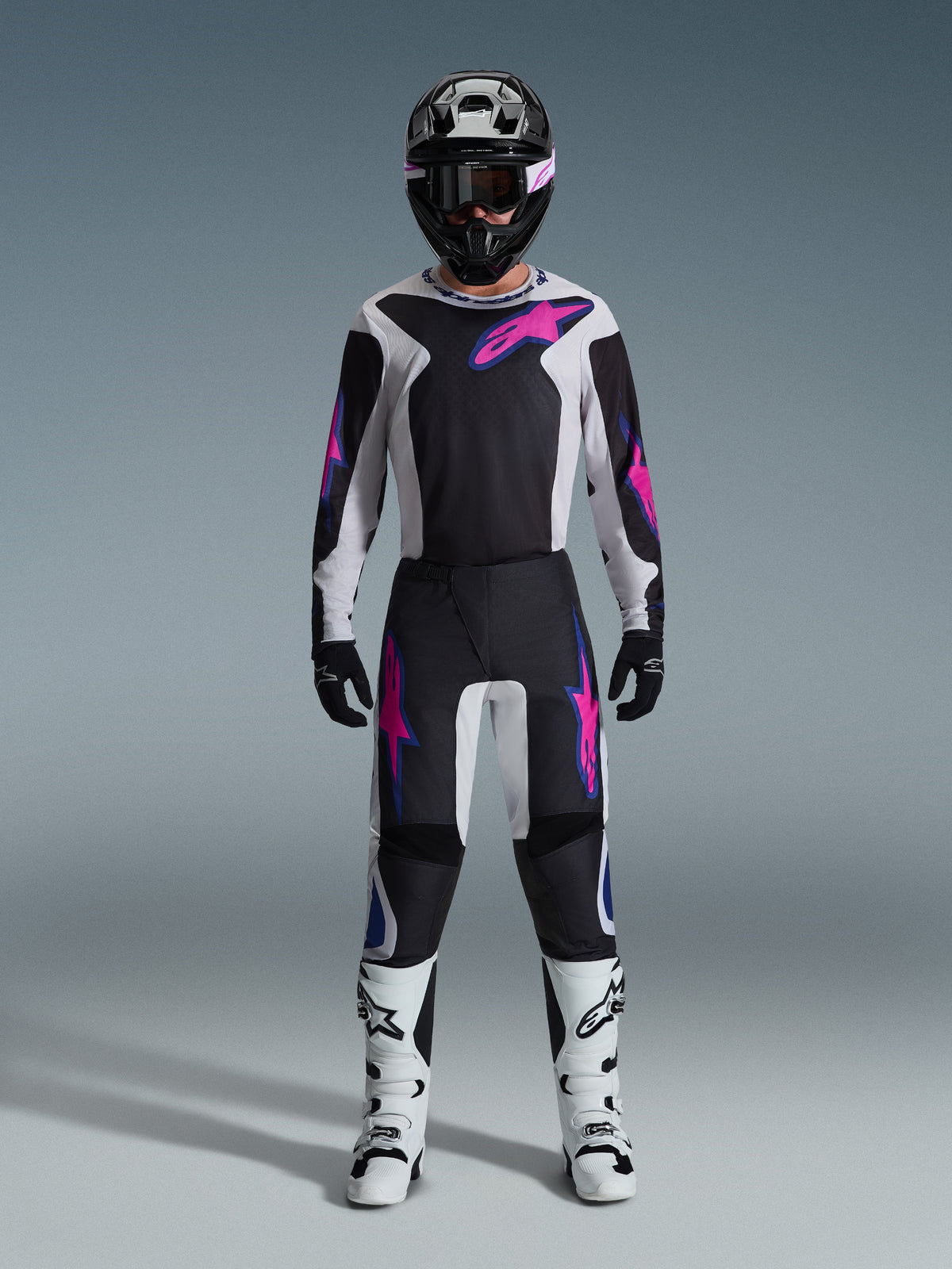 Pilota di motocross con il completo Alpinestars Fluid Grid 2026, maglia nera e grigio chiaro con loghi rosa acceso, pantaloni neri e bianchi con grafiche coordinate rosa e blu, casco nero con maschera rosa, stivali bianchi e neri, equipaggiamento completo per gare off-road