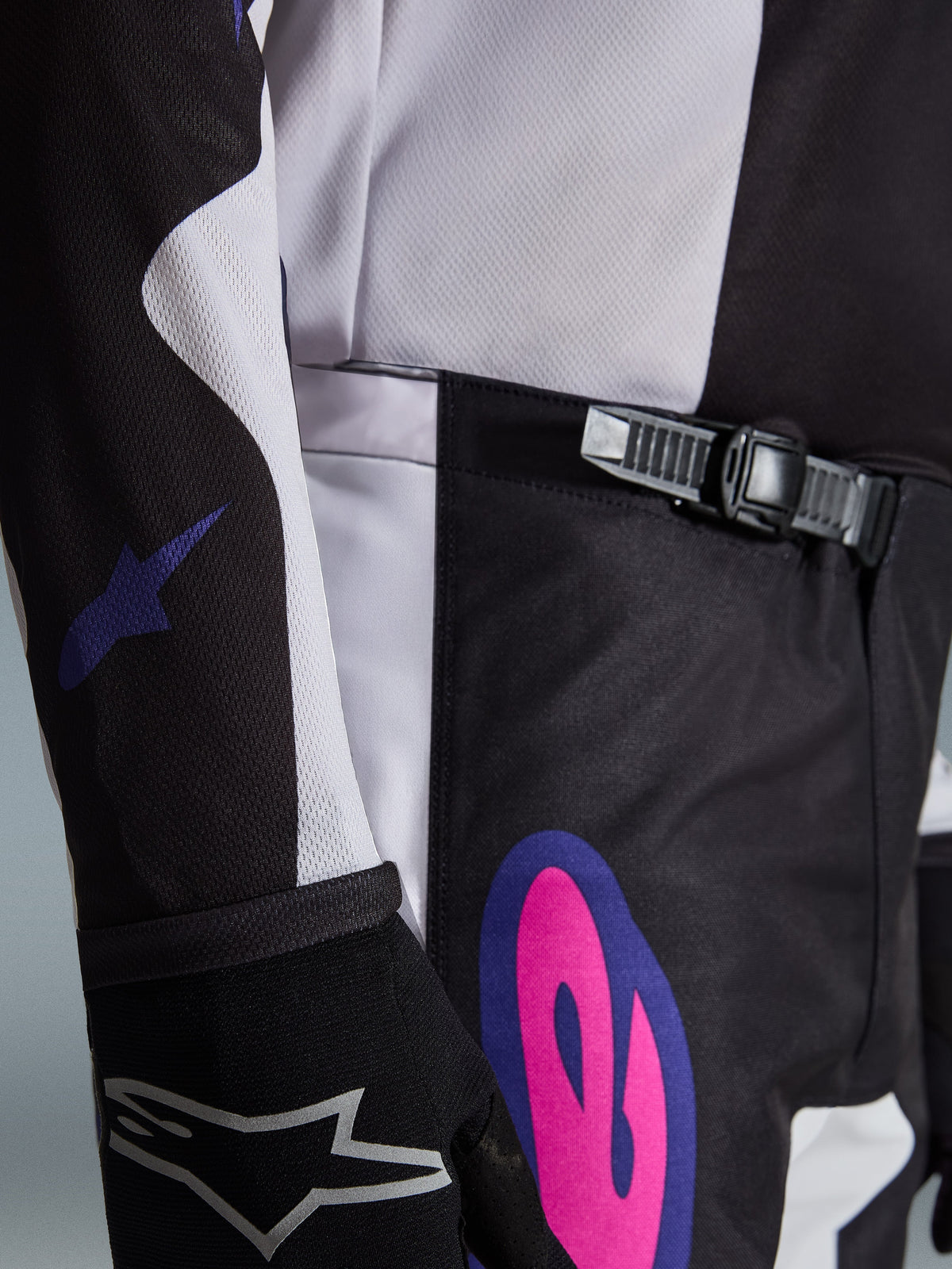 Alpinestars 2026 Fluid Grid Pants, pantaloni da motocross, nero, grigio chiaro e viola, primo piano del sistema di chiusura a cricchetto in vita e di una vivace grafica rosa e viola sul fianco, indossati con maglia coordinata e guanto nero.