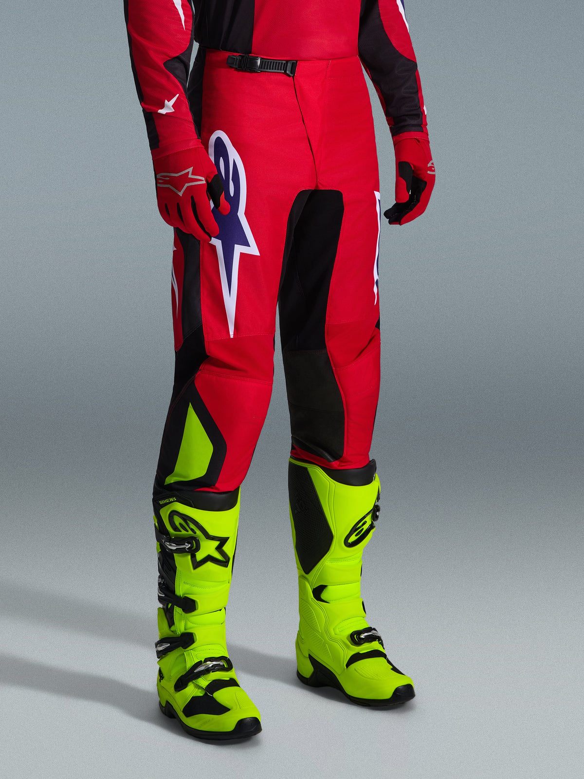 Alpinestars 2026 Fluid Grid Pants, pantaloni da motocross, rosso, nero e viola, indossati con stivali giallo neon e guanti rossi, con pannelli neri e un grande logo a stella viola e bianco, design sport-fit resistente per corse fuoristrada