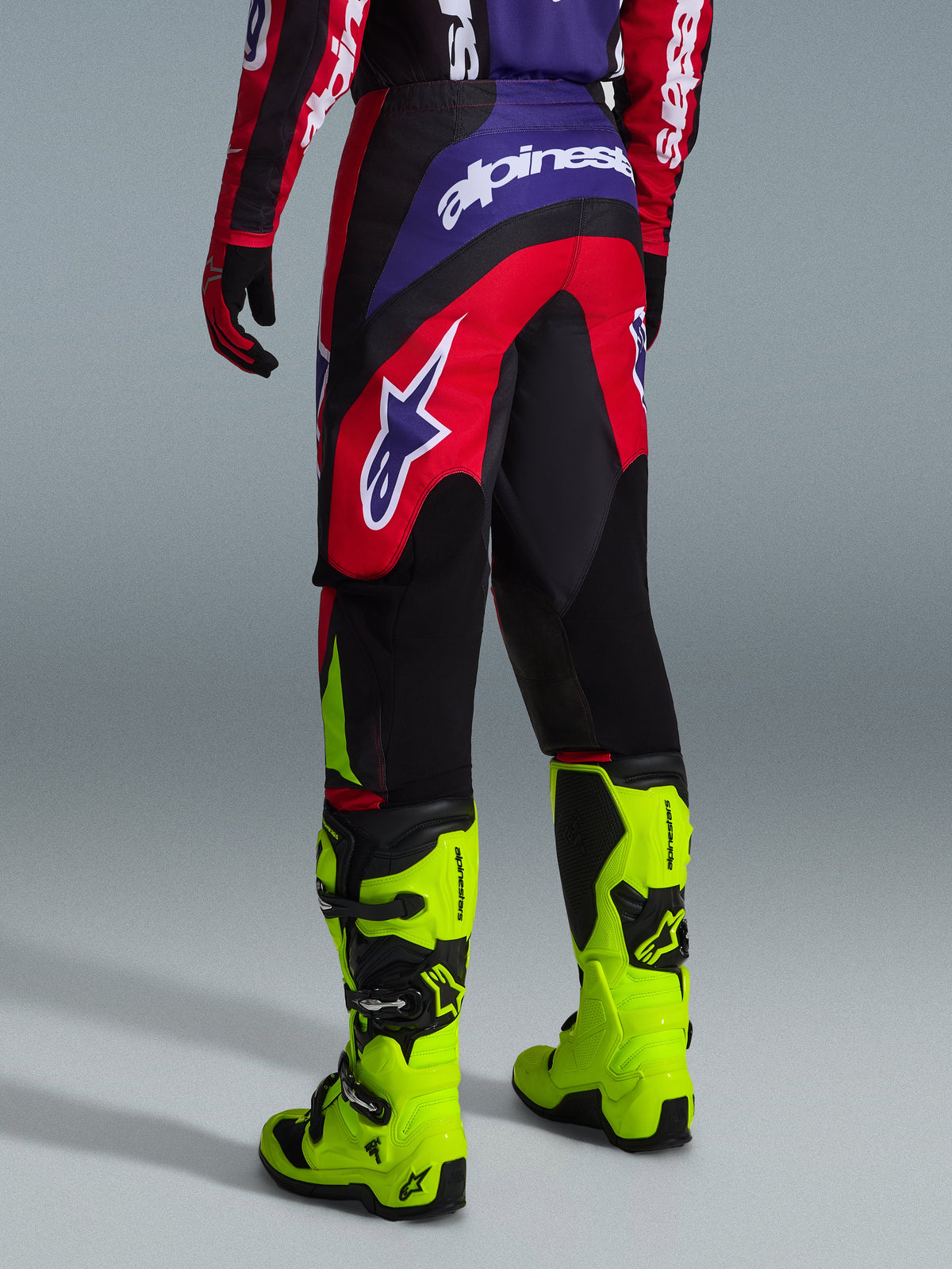 Alpinestars 2026 Fluid Grid Pants, pantaloni da motocross, rosso, nero e viola, vista posteriore con grande logo bianco, indossati con stivali da gara giallo neon e maglia abbinata, design sport-fit resistente per prestazioni fuoristrada
