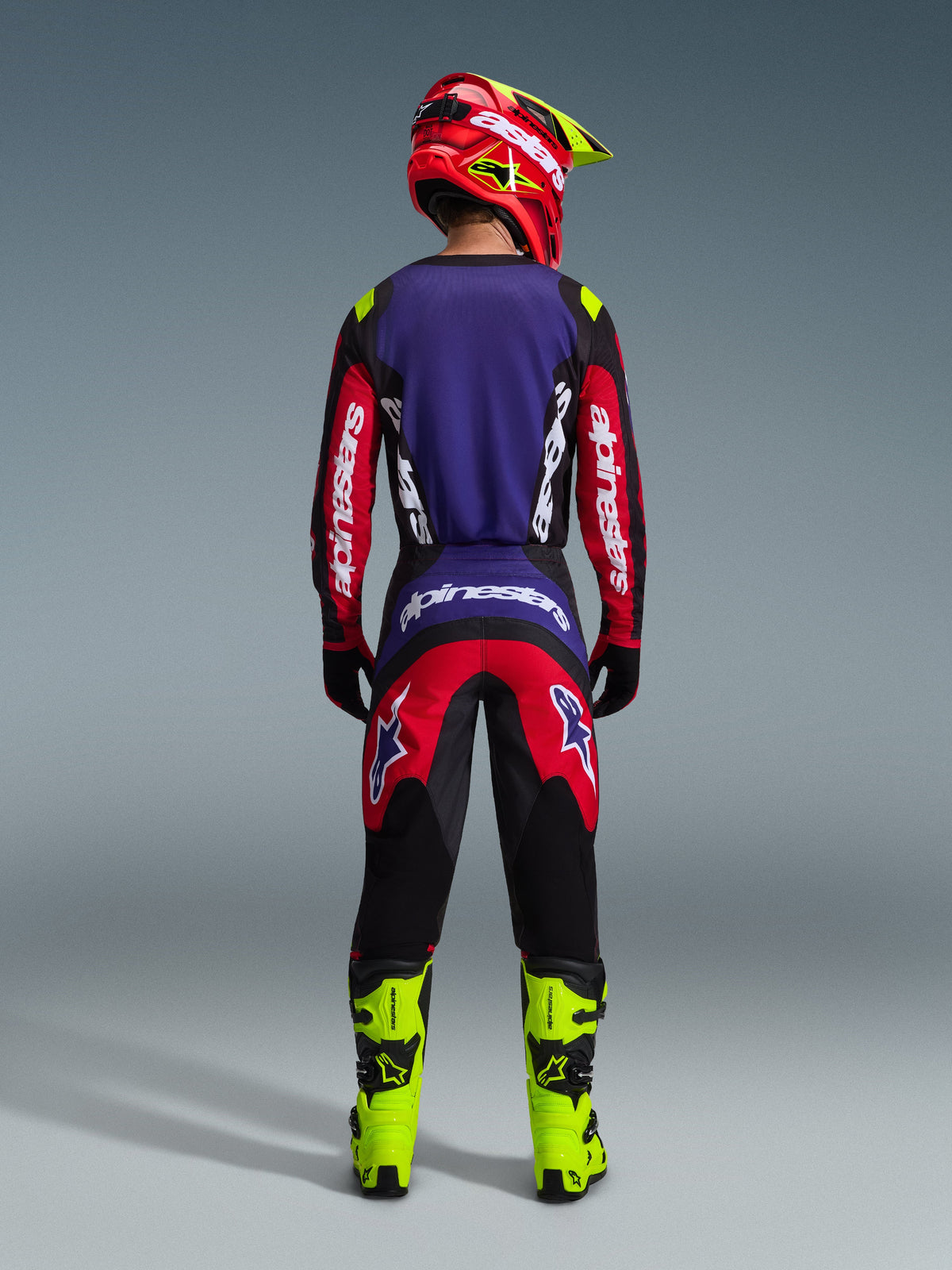 Pilota di motocross visto da dietro con pantaloni Alpinestars 2026 Fluid Grid in rosso, nero e viola, coordinati con maglia abbinata, casco rosso e stivali giallo neon brillante; abbigliamento tecnico da fuoristrada con pannelli rinforzati e design ergonomico, sfondo studio.