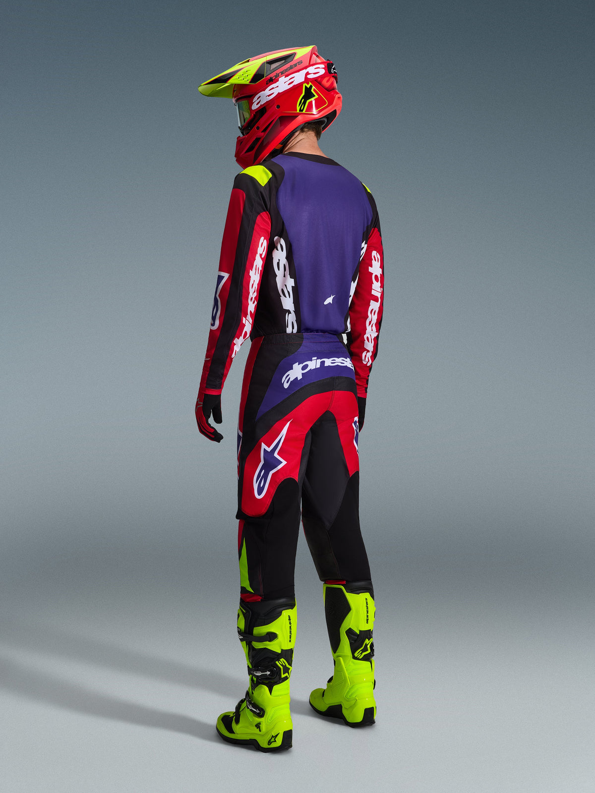 Alpinestars 2026 Fluid Grid Pants, pantaloni da motocross, rosso, nero e viola, indossati da un pilota visto da dietro con maglia coordinata e stivali giallo fluo, con branding audace e design ergonomico per prestazioni off-road.