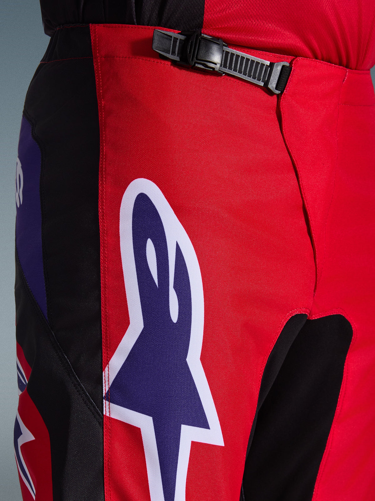 Pantaloni Alpinestars Fluid Grid 2026, pantaloni da motocross, rosso nero e viola, primo piano della zona della vita e dei fianchi con sistema di chiusura a cricchetto nero e grande logo Alpinestars a stella viola con bordo bianco, tessuto strutturato resistente per corse off-road