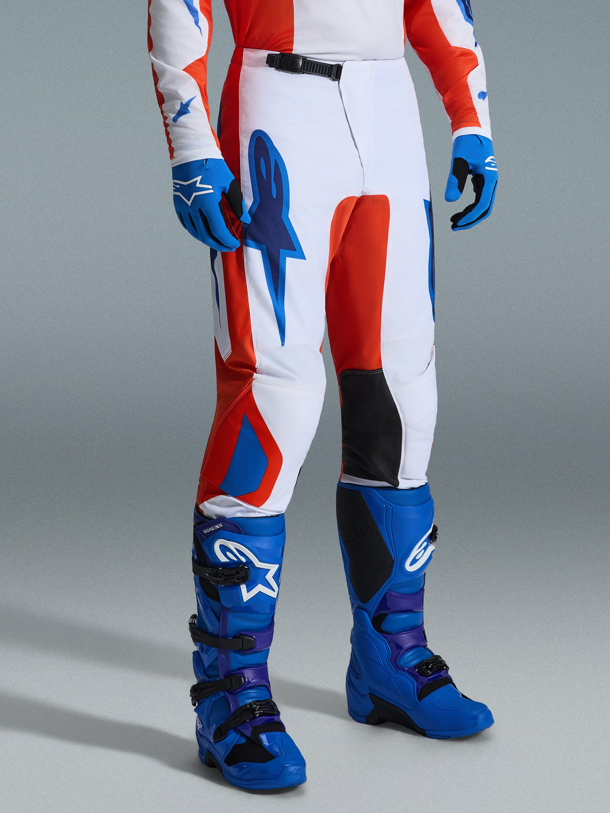 Alpinestars 2026 Fluid Grid Pants, pantaloni da motocross, arancione e blu UCLA su base bianca, indossati con guanti e stivali blu coordinati, grafiche del brand, pannelli interni neri sulle ginocchia e design ergonomico per la guida fuoristrada.
