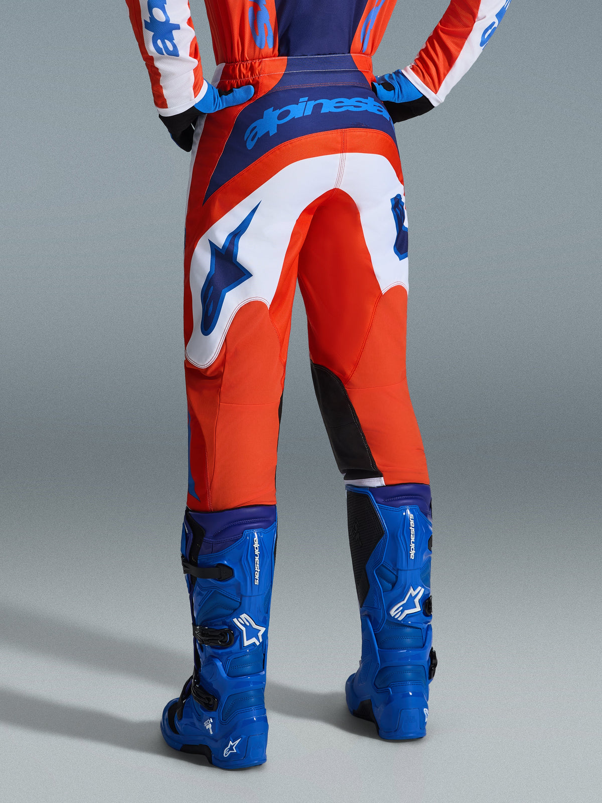 Alpinestars 2026 Fluid Grid Pants, pantaloni da motocross, arancione e blu UCLA con pannelli bianchi, vista posteriore con logo blu sulla seduta e logo a stella, abbinati a stivali da motocross blu coordinati, equipaggiamento professionale per fuoristrada