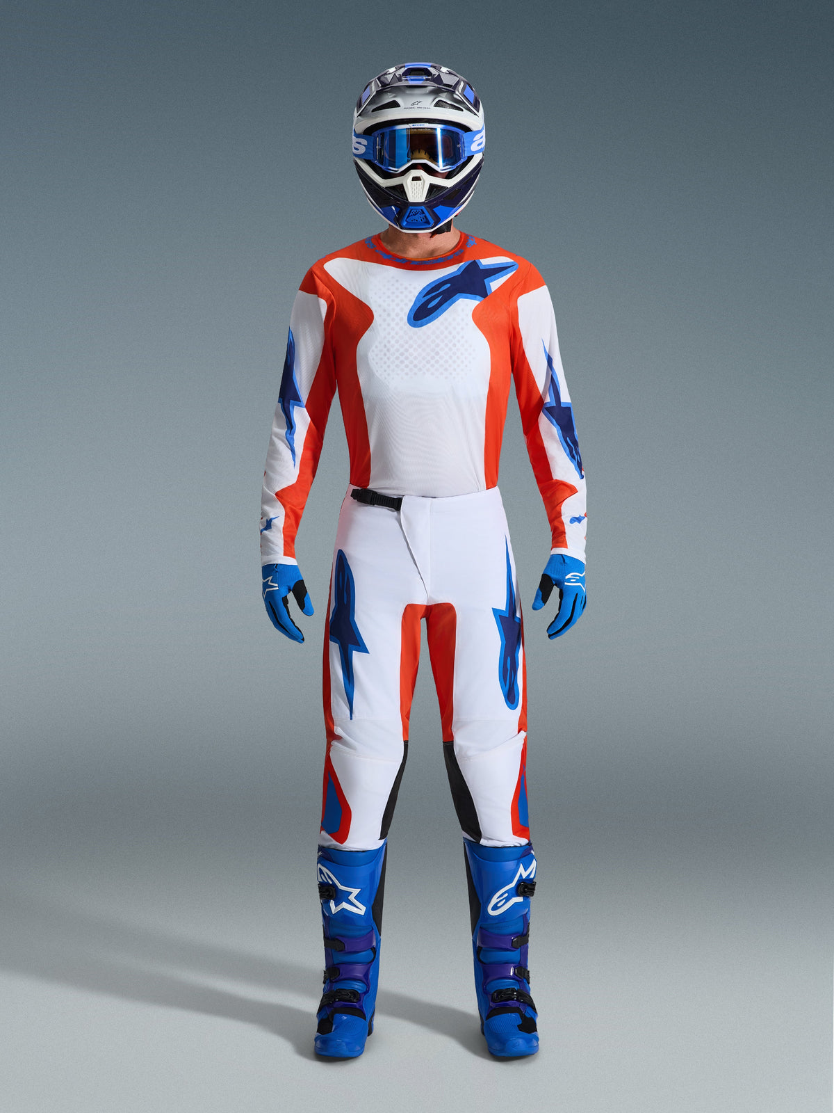 Pilota di motocross con abbigliamento completo da gara, pantaloni Alpinestars 2026 Fluid Grid in arancione e blu UCLA, maglia bianca con maniche arancioni e loghi blu, casco e occhiali blu, stivali e guanti blu, completo coordinato per corse fuoristrada