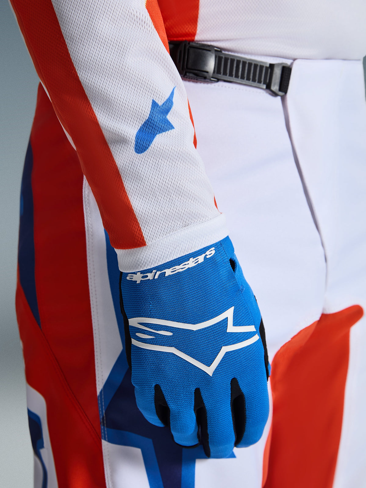 Alpinestars 2026 Fluid Grid Pants, pantaloni da motocross, arancione e blu UCLA, primo piano con guanto blu coordinato e manica della maglia in mesh bianca con loghi blu, dettaglio della chiusura a cricchetto in vita e vestibilità atletica