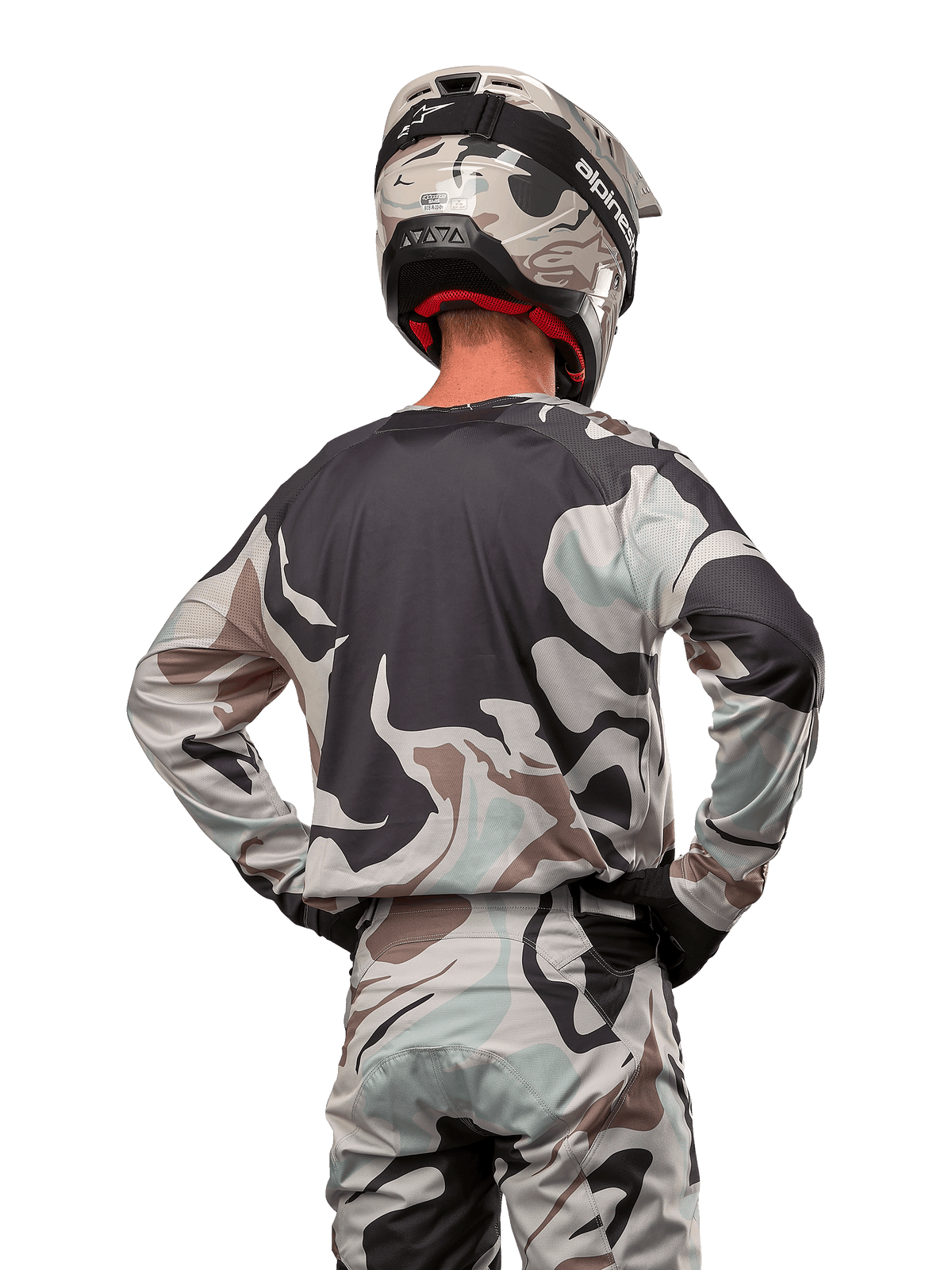 Pilota di motocross visto da dietro che indossa la maglia Alpinestars Racer Tactical 2024 in Iron Camo Dust Gray, pantaloni e casco mimetici coordinati, mani sui fianchi, abbigliamento da gara fuoristrada coordinato.