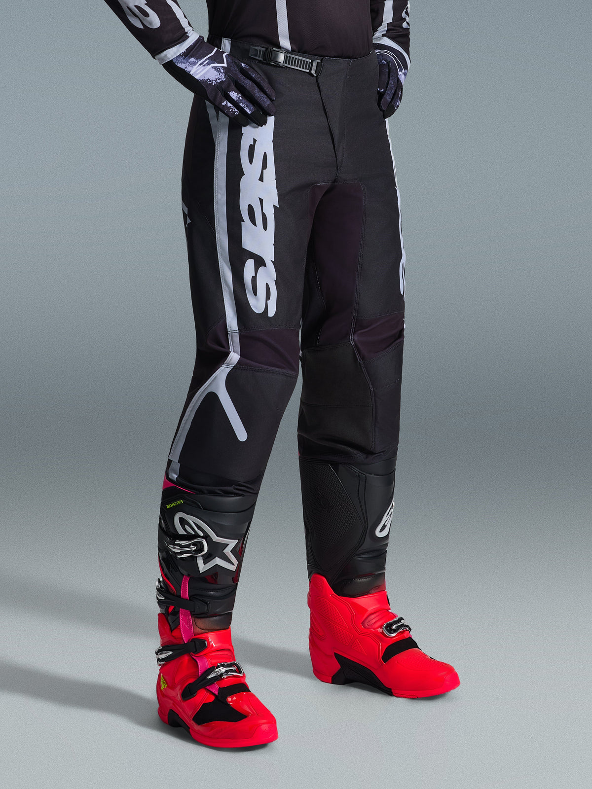 Pantaloni Alpinestars 2026 Fluid Apex, pantaloni da motocross, nero e grigio, indossati con stivali rosa vivace e rossi e maglia abbinata nera e bianca, con logo Alpinestars verticale sulla coscia, pannelli rinforzati e chiusura a cricchetto in vita per prestazioni fuoristrada.