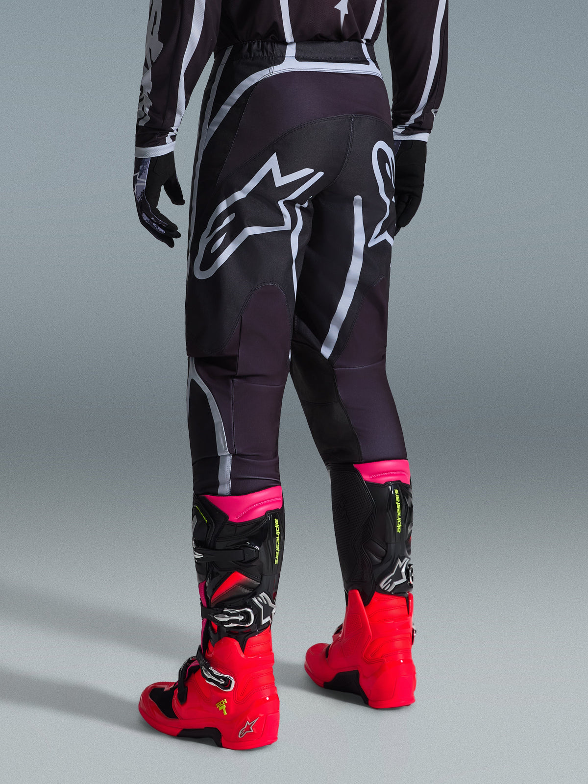 Alpinestars Pantaloni Fluid Apex 2026, pantaloni da motocross, nero e grigio, vista posteriore con grandi loghi Alpinestars bianchi sulla seduta, abbinati a stivali rosso acceso e rosa, costruzione in poliestere 600D per prestazioni off-road