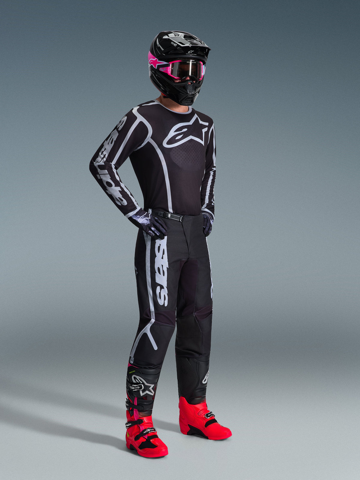 Pantaloni Alpinestars Fluid Apex 2026, pantaloni da motocross, nero e grigio, indossati con maglia coordinata nera e bianca, casco nero con maschera rosa e stivali rosso acceso, con vistoso branding bianco sulle gambe e design sport-fit per il fuoristrada.