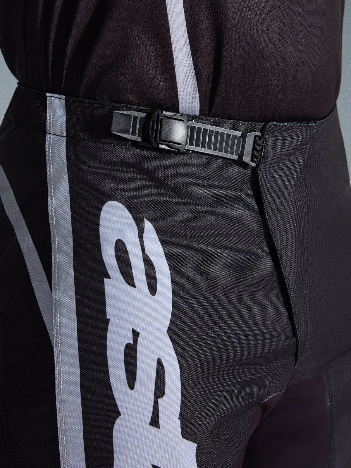Alpinestars 2026 Fluid Apex Pants, pantaloni da motocross, nero e grigio, dettaglio ravvicinato del sistema di chiusura a cricchetto integrato per una vestibilità sicura, con grafiche del logo bianche su resistente politessuto 600D.