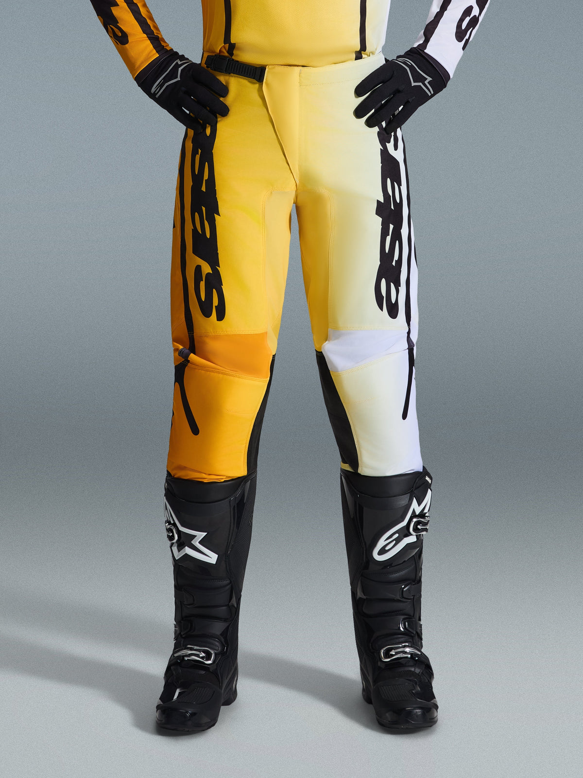 Alpinestars 2026 Fluid Apex Pants, pantaloni da motocross, giallo e nero, design bicolore con pannelli gialli e bianchi, vistoso logo 'astars' verticale, indossati con stivali e guanti da motocross neri, abbigliamento tecnico per guida fuoristrada
