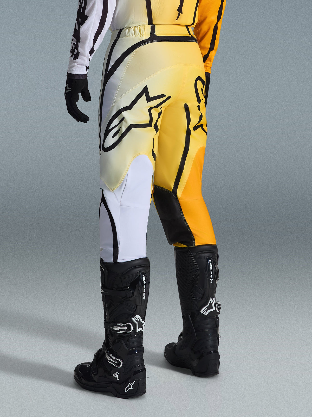 Alpinestars 2026 Fluid Apex Pants, pantaloni da motocross, sfumatura giallo e nero con accenti bianchi e arancioni, vista posteriore, indossati con maglia coordinata e stivali Tech 7 neri, con evidente logo Alpinestars nero sulla seduta, abbigliamento tecnico per fuoristrada