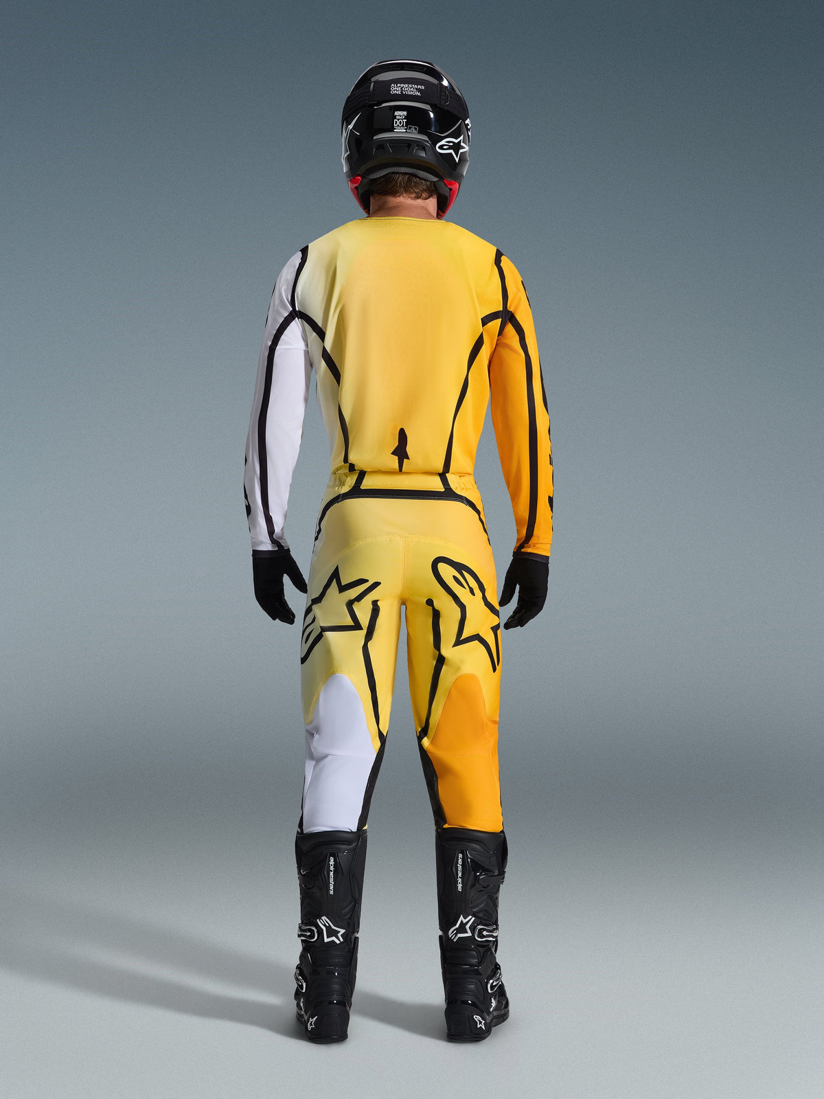 Alpinestars Pantaloni Fluid Apex 2026, pantaloni da motocross, giallo e nero, vista posteriore di un pilota con equipaggiamento completo da motocross, maglia abbinata con sfumature bianche e arancioni, casco nero, stivali neri e guanti neri, completo coordinato da fuoristrada