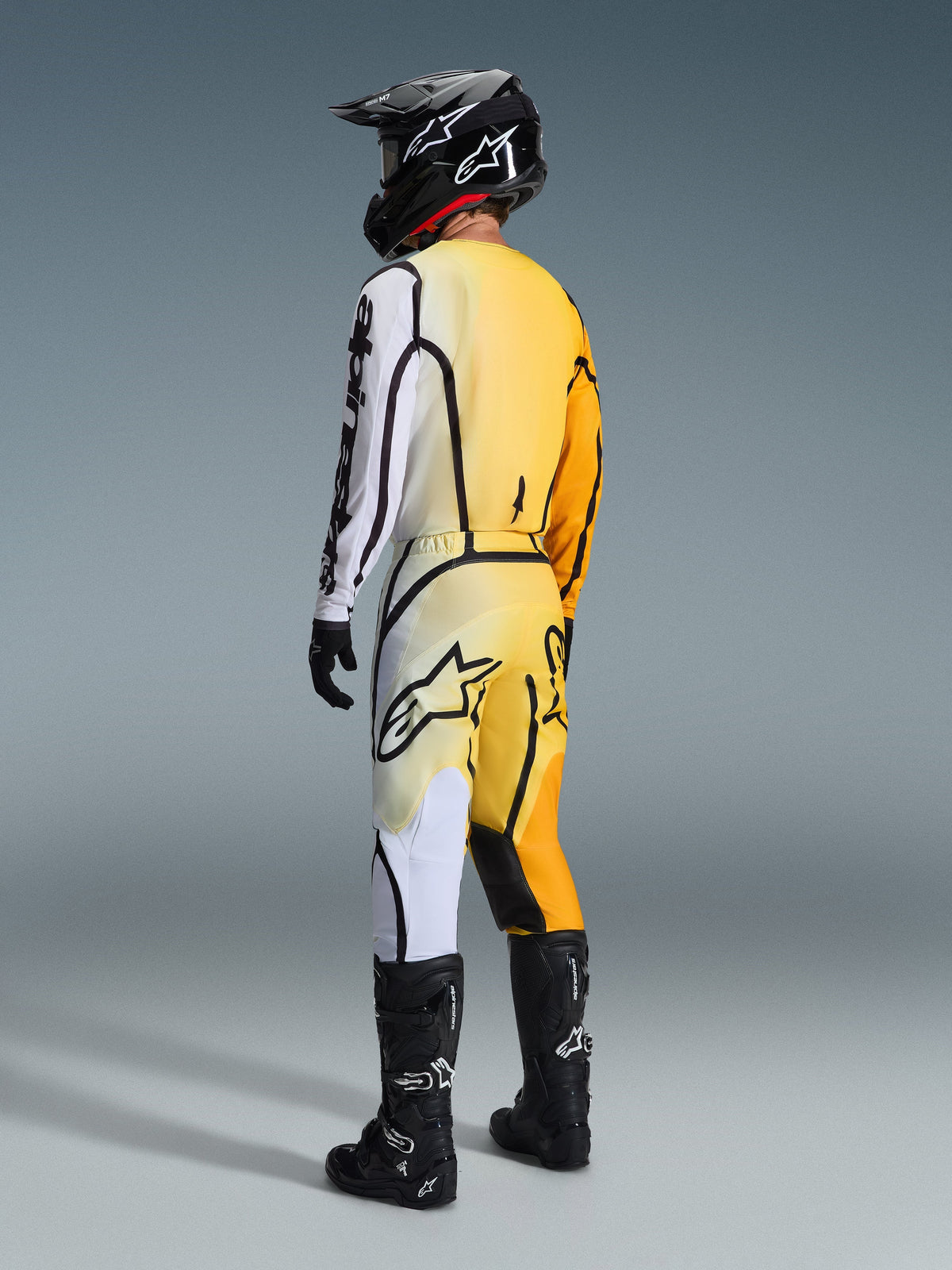 Pantaloni Alpinestars Fluid Apex 2026, pantaloni da motocross, sfumatura giallo e bianco con accenti neri, vista posteriore del pilota con equipaggiamento da gara completo che include maglia abbinata, casco nero e stivali neri