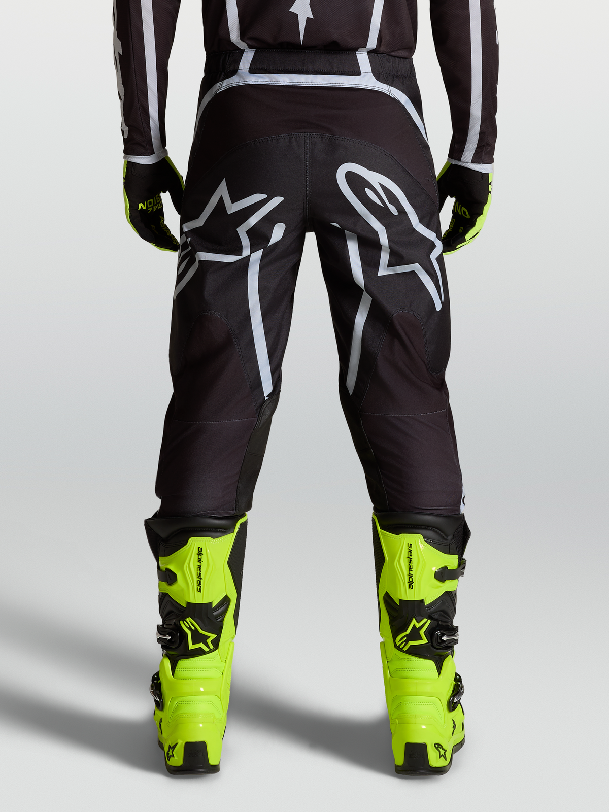 Alpinestars Pantaloni Fluid Apex V2, pantaloni motocross da uomo, nero e argento, vista posteriore con grandi loghi Astars e dettagli in maglia mesh, indossati con stivali giallo fluo per performance fuoristrada