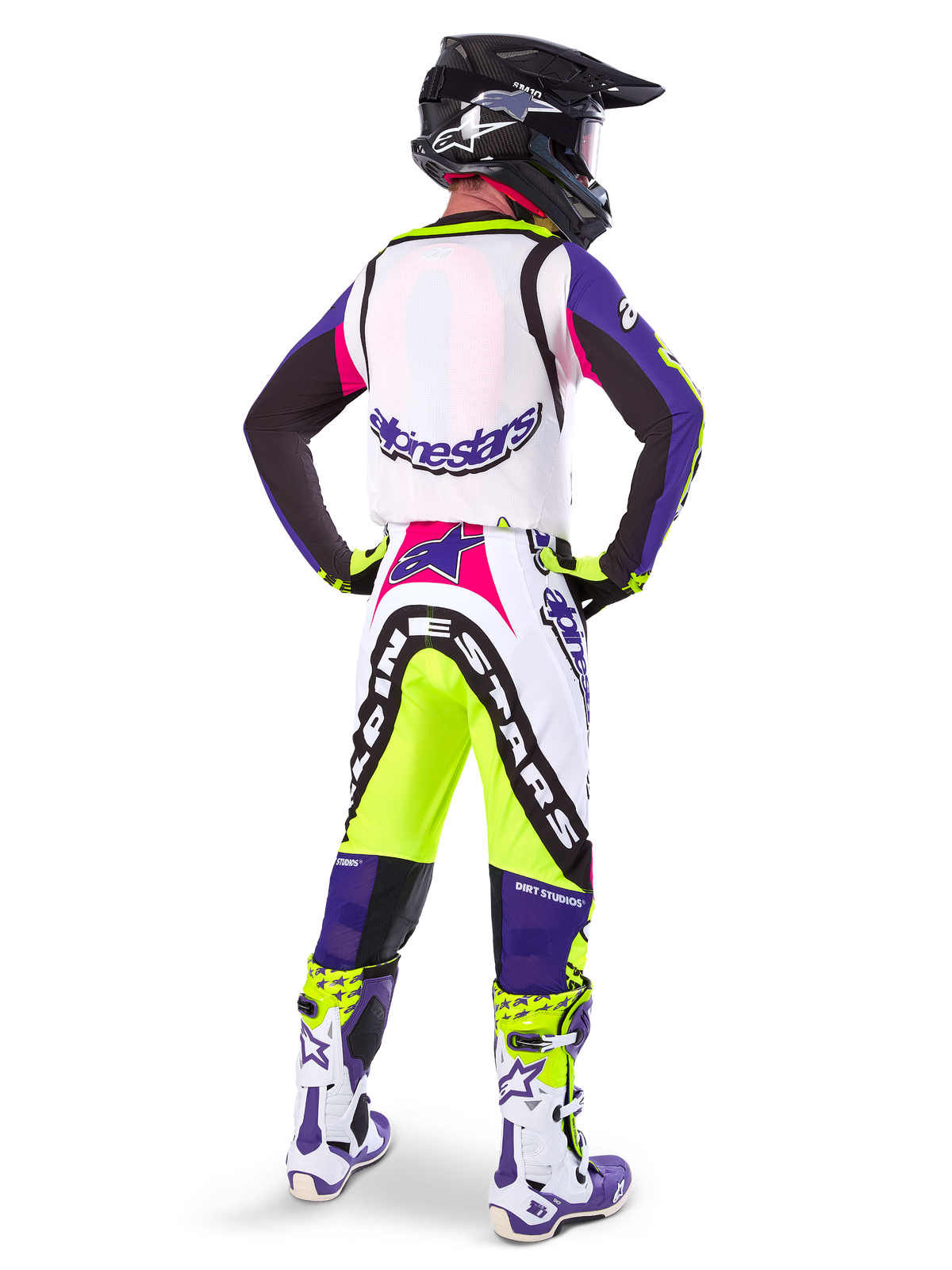 Pilota di motocross che indossa pantaloni Alpinestars Limited Edition Supertech Dirt Studios® in Bianco, Viola e Giallo Fluo, visto da dietro con maglia e stivali coordinati e casco in carbonio nero, abbigliamento professionale da gara off-road con grafiche audaci e accenti neon.