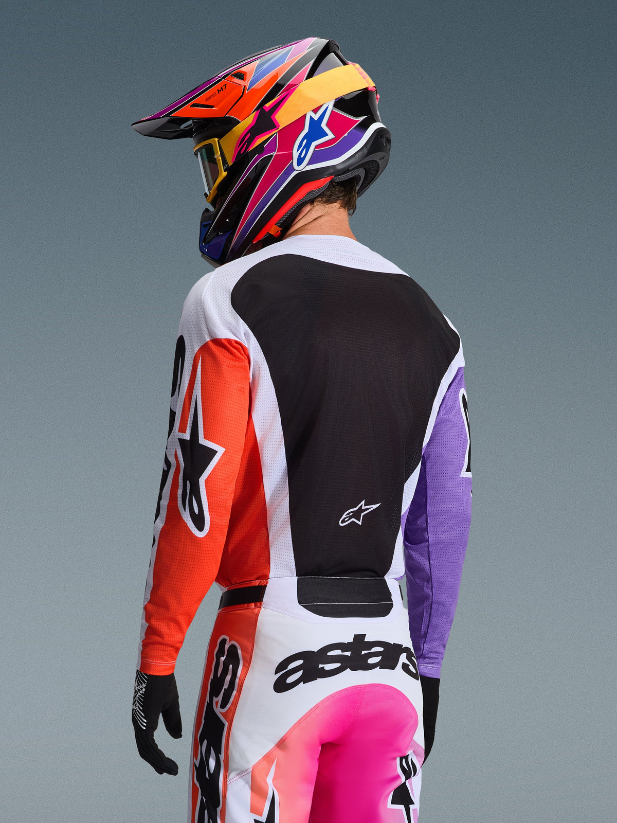 Pilota di motocross visto da dietro che indossa la maglia Alpinestars 2026 Racer Air Portl in Multicolor, caratterizzata da manica destra arancione, manica sinistra viola e un ampio pannello posteriore in mesh nero per la ventilazione, abbinata a pantaloni da motocross coordinati e un vivace casco multicolore, equipaggiamento professionale per corse fuoristrada
