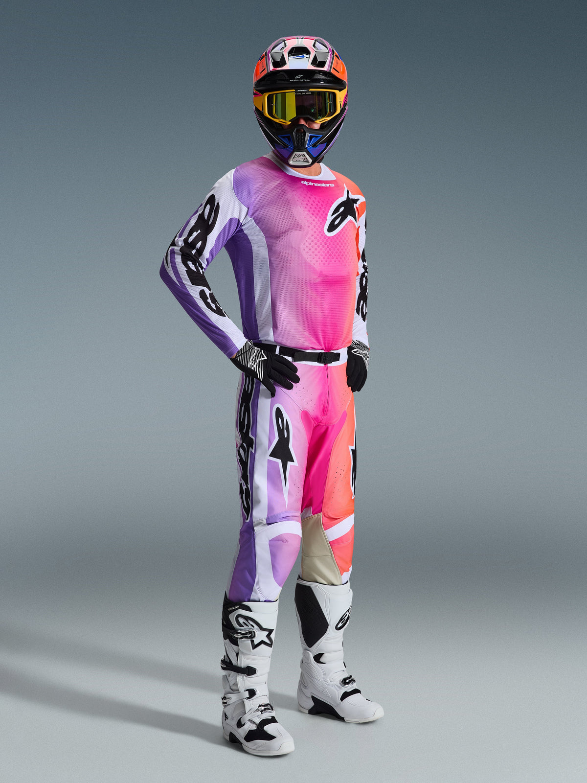 Pilota di motocross con maglia Alpinestars Racer Air Portl 2026 e pantaloni coordinati, sfumatura multicolore rosa, viola e arancione con accenti bianchi, casco colorato e maschera gialla coordinati, stivali bianchi e neri, completo da gara per prestazioni off-road