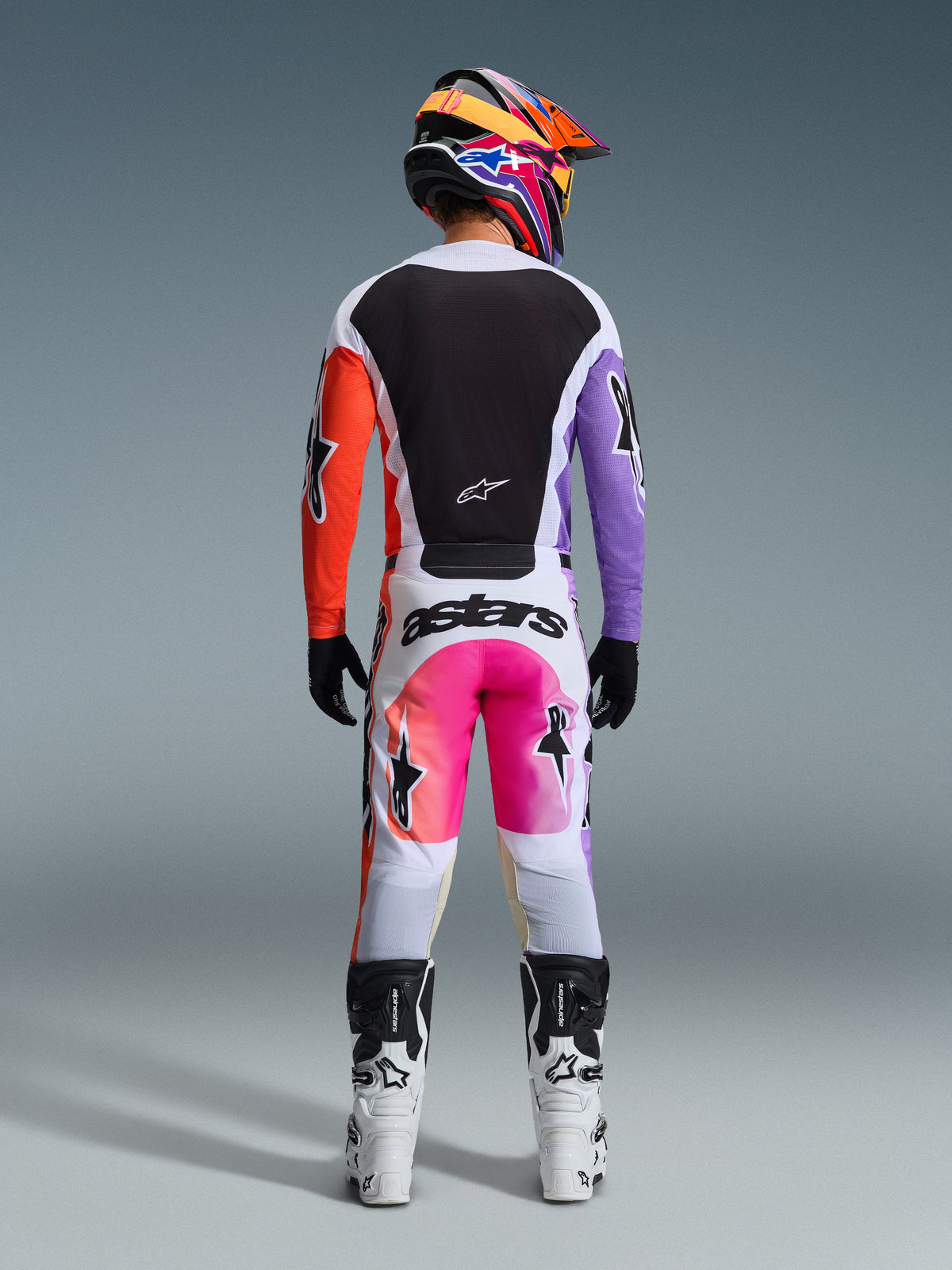 Pilota di motocross con abbigliamento completo da gara visto dal retro, maglia Alpinestars 2026 Racer Air Portl con pannelli bianchi, neri, arancioni e viola, pantaloni bianchi coordinati con dettagli rosa e marchio 'astars', stivali bianchi e neri e casco vivace multicolore, completo coordinato per corse di motocross
