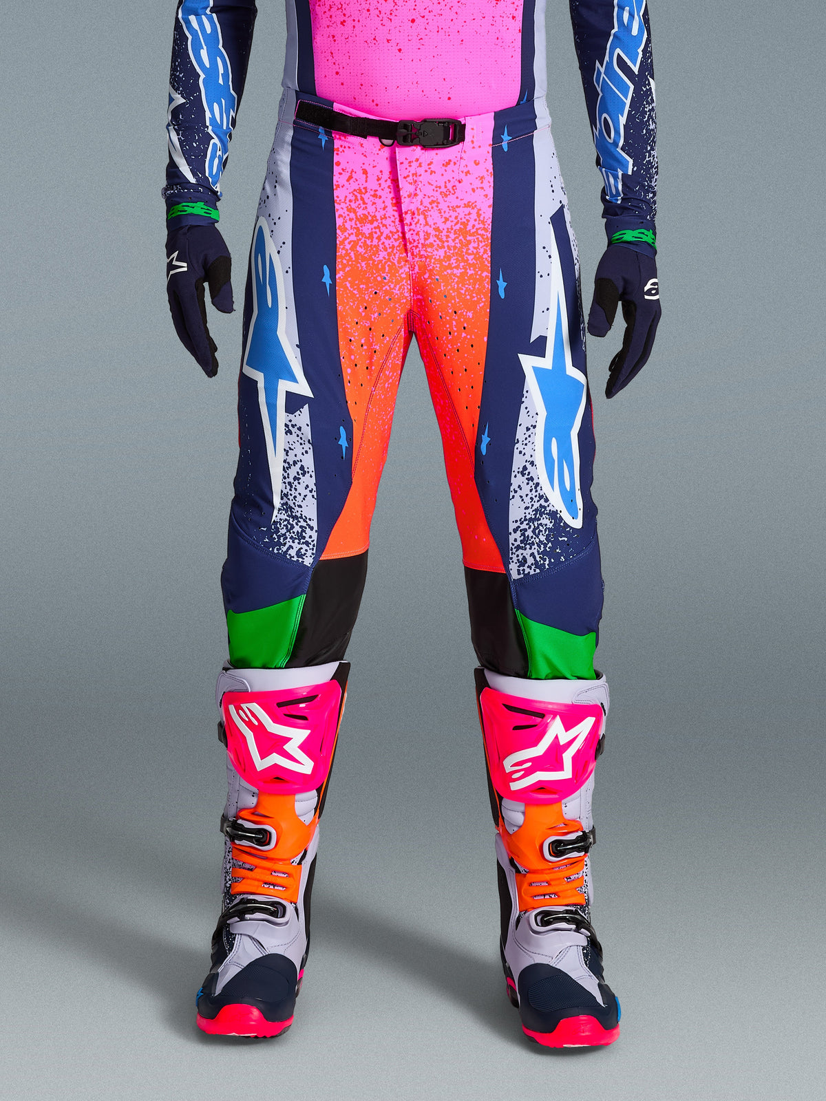 Alpinestars Limited Edition Supertech Records Pants, pantaloni da motocross, multicolore con vivaci grafiche splash rosa e arancione, pannelli blu navy con loghi a stella blu e accenti verdi, indossati con maglia coordinata e stivali da motocross grigi, rosa e arancioni.