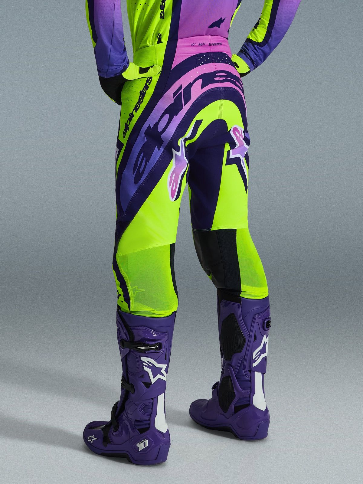 Alpinestars 2026 Supertech Vista Pants, pantaloni da motocross, giallo fluo, viola e nero, vista posteriore indossata con maglia coordinata e stivali da motocross viola, con branding audace e zone in mesh traspirante sui polpacci