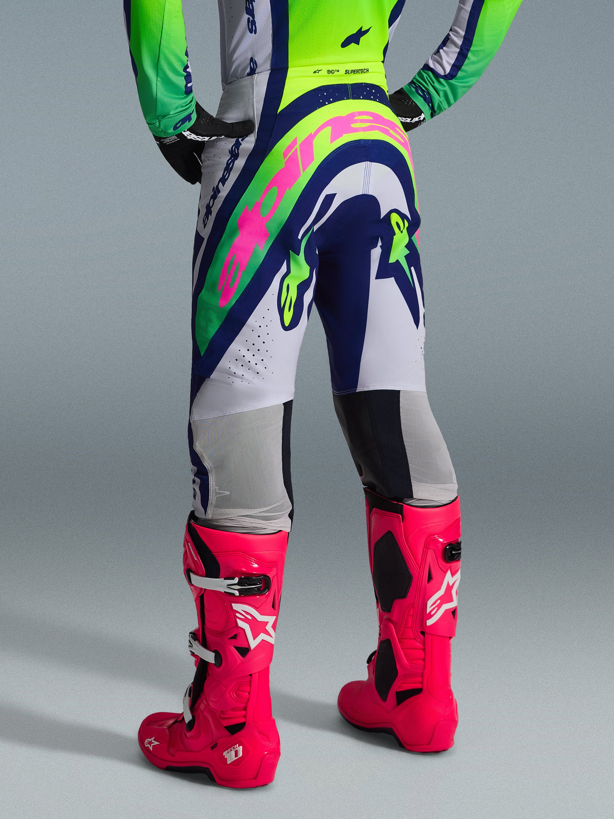 Alpinestars 2026 Supertech Vista Gear Set, vista posteriore dell'abbigliamento da motocross, pantaloni bianchi con pannelli blu e verde lime, grande logo rosa sulla seduta, abbinati a vivaci stivali Tech 10 rosa, equipaggiamento professionale da gara off-road