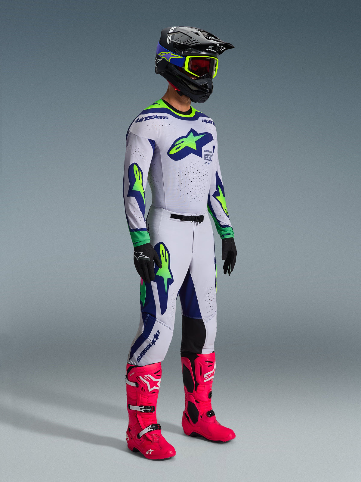 Alpinestars 2026 Supertech Vista Gear Set, pilota di motocross in abbigliamento da gara completo, maglia e pantaloni bianchi con dettagli verde lime e blu navy, stivali rosa acceso, casco nero con dettagli blu e lime, abbigliamento off-road coordinato di livello professionale.