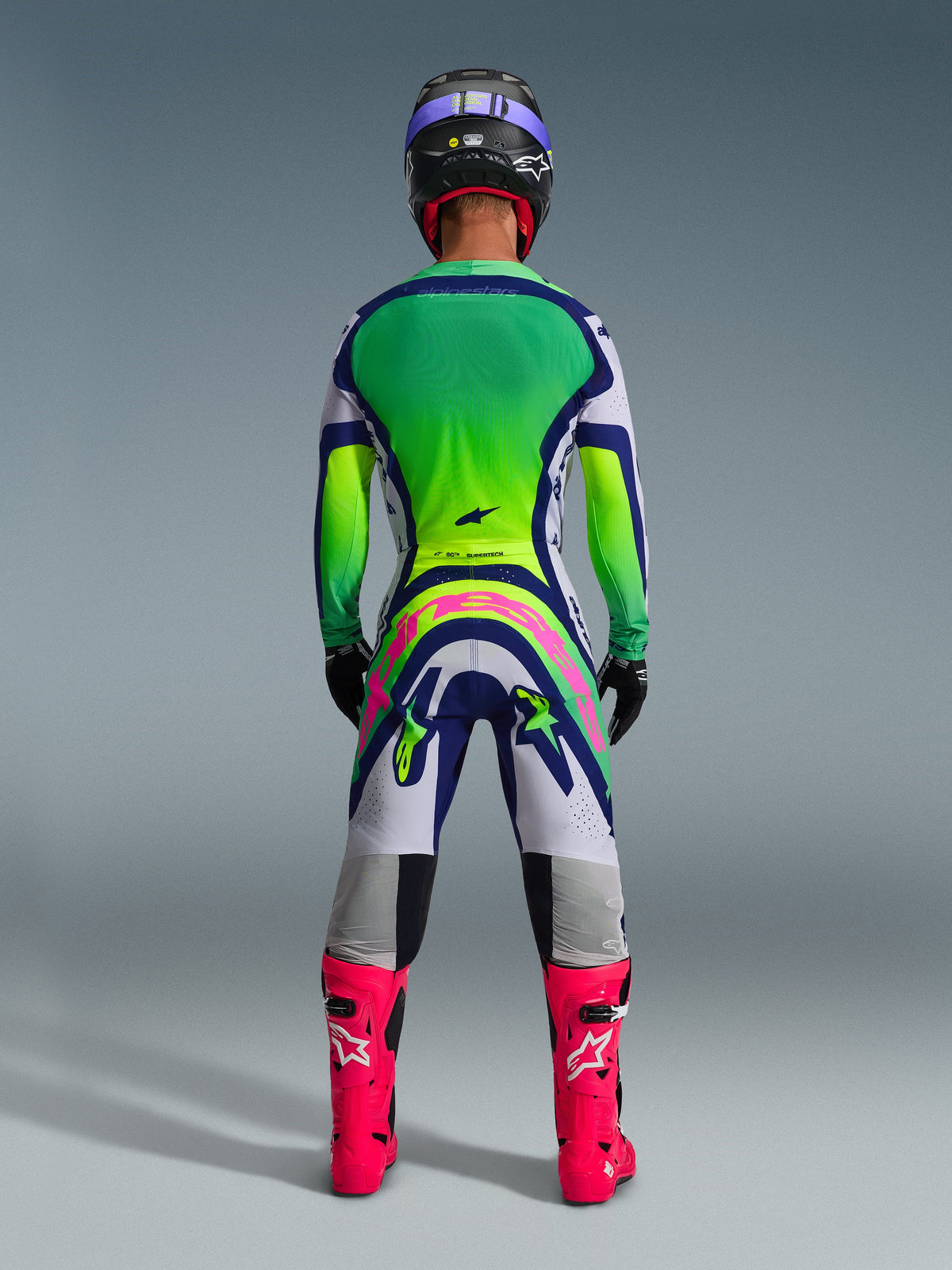 Alpinestars 2026 Supertech Vista Gear Set, vista posteriore del completo da motocross, maglia con sfumatura verde e gialla, pantaloni bianchi e blu con grande logo rosa sul retro, stivali rosa acceso e casco viola, abbigliamento professionale per gare fuoristrada