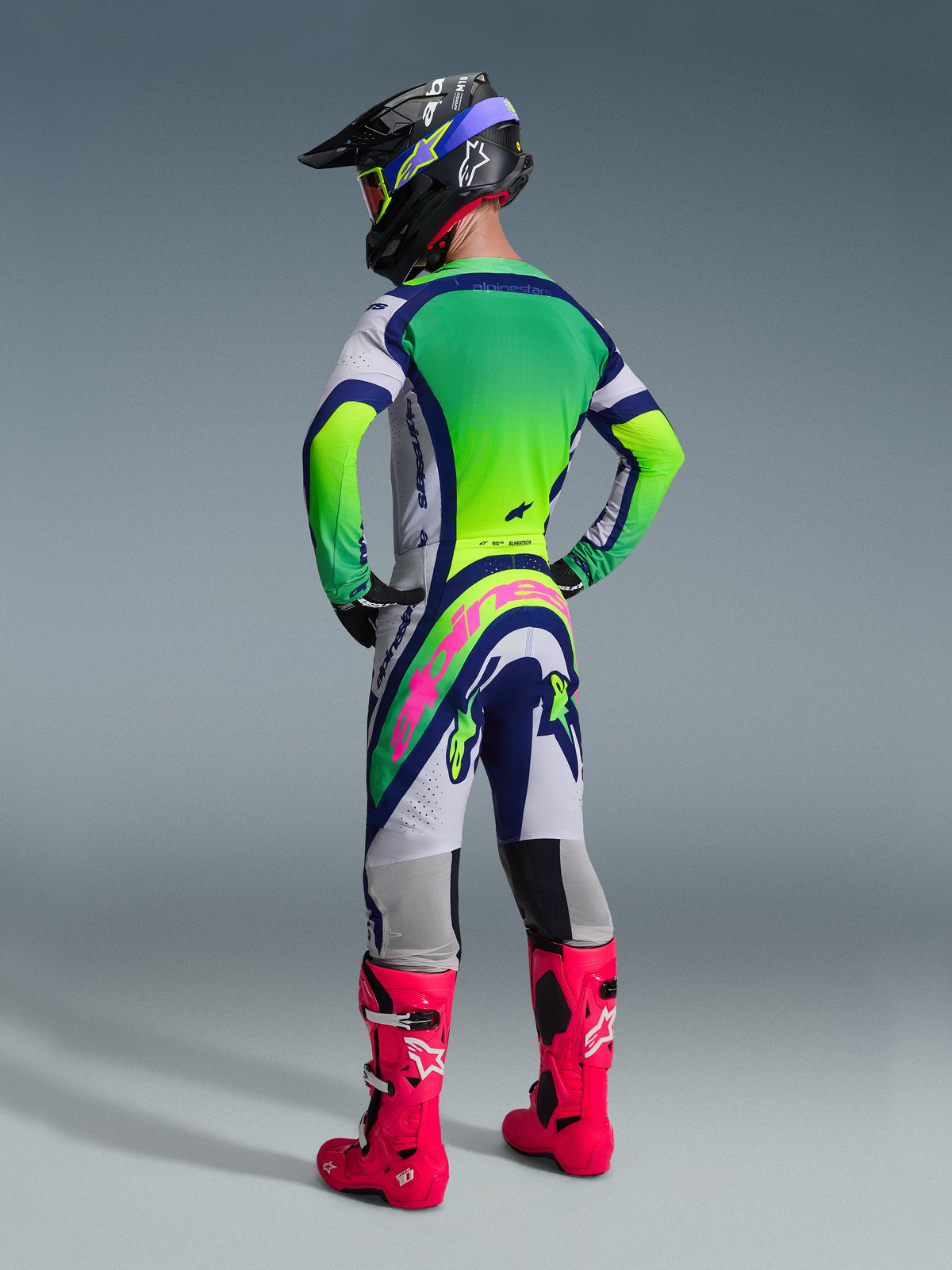 Vista posteriore di un pilota di motocross che indossa il completo Alpinestars 2026 Supertech Vista, maglia bianca e verde lime con accenti blu, pantaloni coordinati con un vistoso logo rosa sul retro, stivali da corsa rosa acceso e un casco nero con occhiali verde lime, abbigliamento professionale per gare off-road