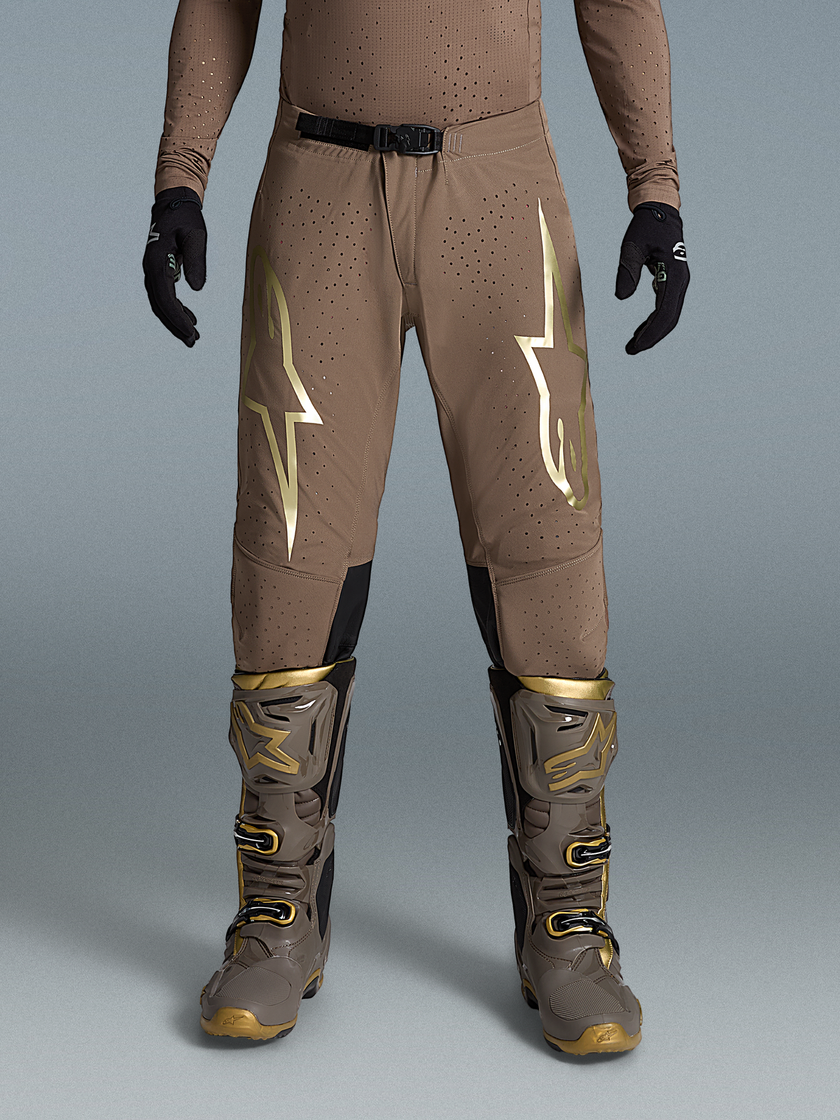 Alpinestars Pantaloni Supertech Air Gold Edizione Limitata, pantaloni da motocross marrone e oro, tessuto elastico traspirante traforato con loghi dorati, indossati con stivali e guanti coordinati.