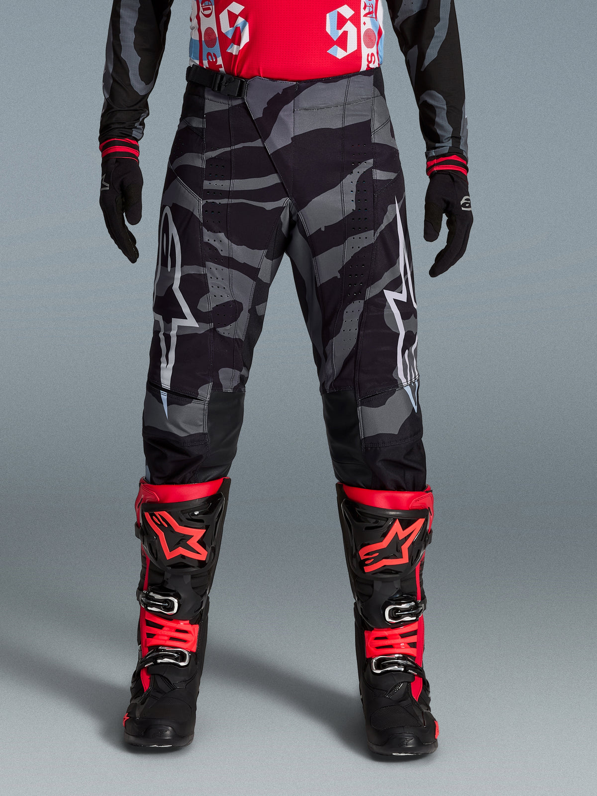 Alpinestars Pantaloni Techstar Beast Coast edizione limitata, pantaloni da motocross con motivo camouflage nero e grigio, indossati con guanti e stivali coordinati neri e arancioni, abbigliamento tecnico per off-road