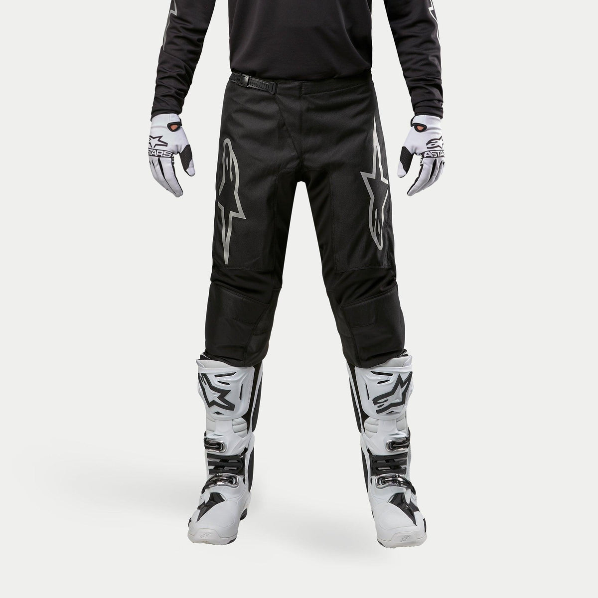 Alpinestars 2024 Fluid Graphite Pants, pantaloni da motocross, nero e argento, con loghi Alpinestars argento sulle cosce, indossati con stivali e guanti da motocross bianchi e neri, design sportivo per corse fuoristrada