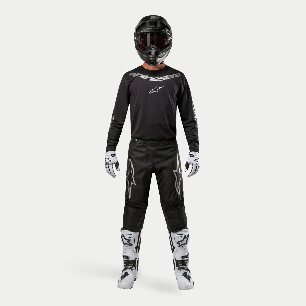 Pilota di motocross con equipaggiamento da gara completo, pantaloni Alpinestars Fluid Graphite 2024 in nero e argento, maglia nera a maniche lunghe con loghi argento, casco nero con occhiali, stivali bianchi e neri, guanti bianchi e neri, completo coordinato da motocross