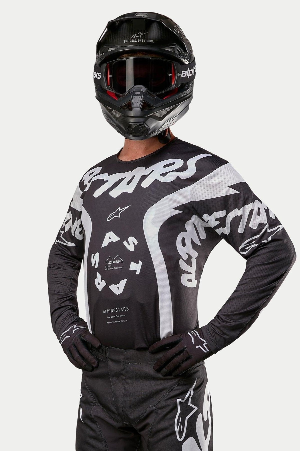 Alpinestars 2024 Racer Hana Jersey, maglia da motocross, nero e bianco, con grafiche testuali e logo Alpinestars in bianco, indossata da un pilota con casco nero e maschera, abbigliamento ad alte prestazioni per corse fuoristrada