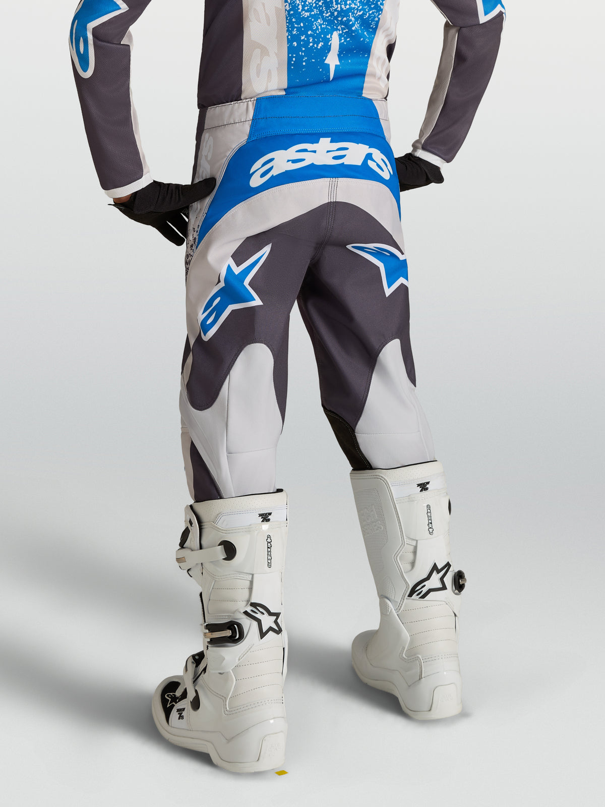 Alpinestars Pantaloni Fluid Drag Ragazzo, pantaloni da motocross grigi e blu, visti da dietro su un pilota con stivali bianchi, abbigliamento tecnico off-road.